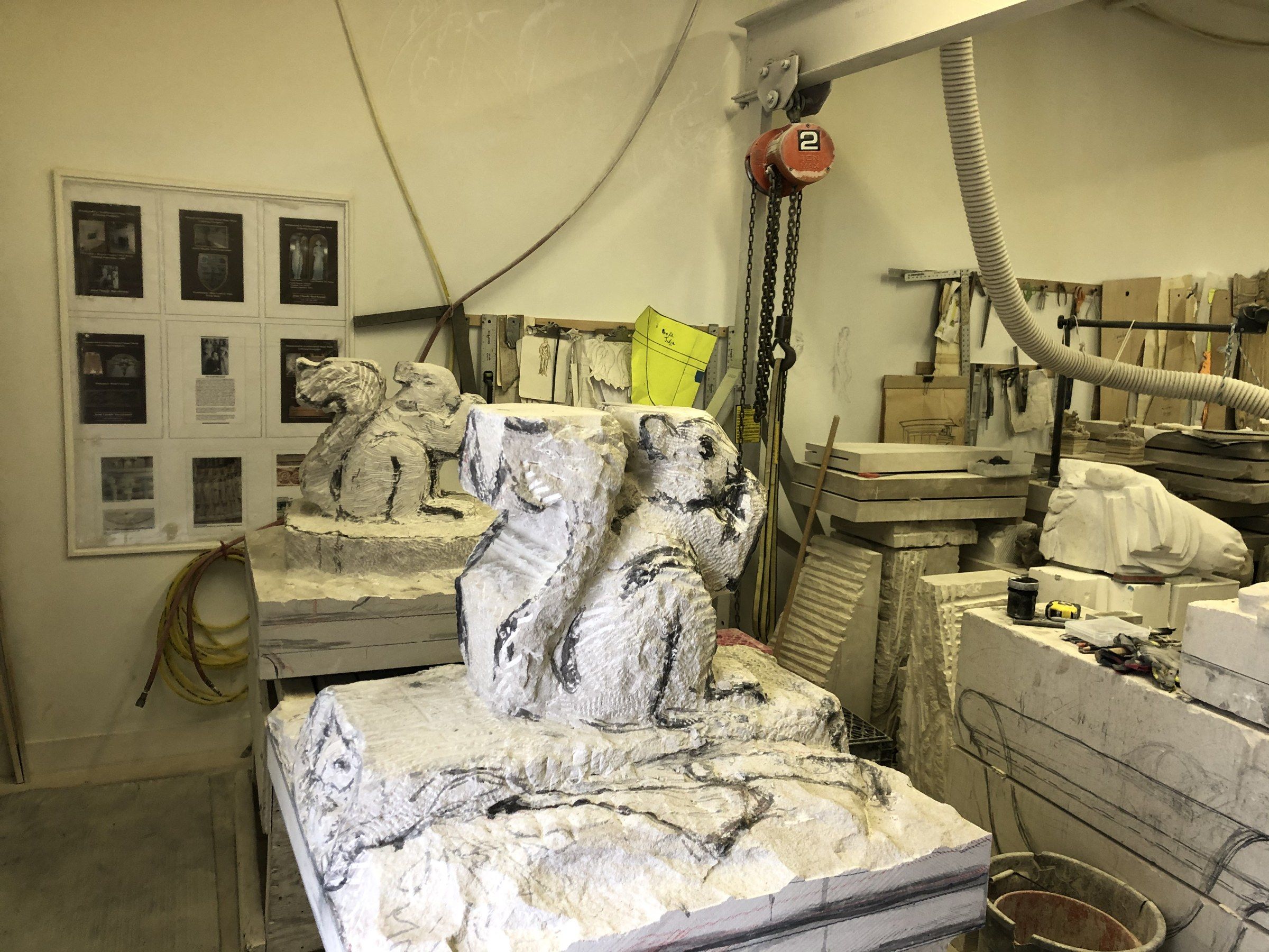 Two squirrels roughed out of their blocks on benches in the studio, the nearer one covered in charcoal guide lines above a rough, square base.