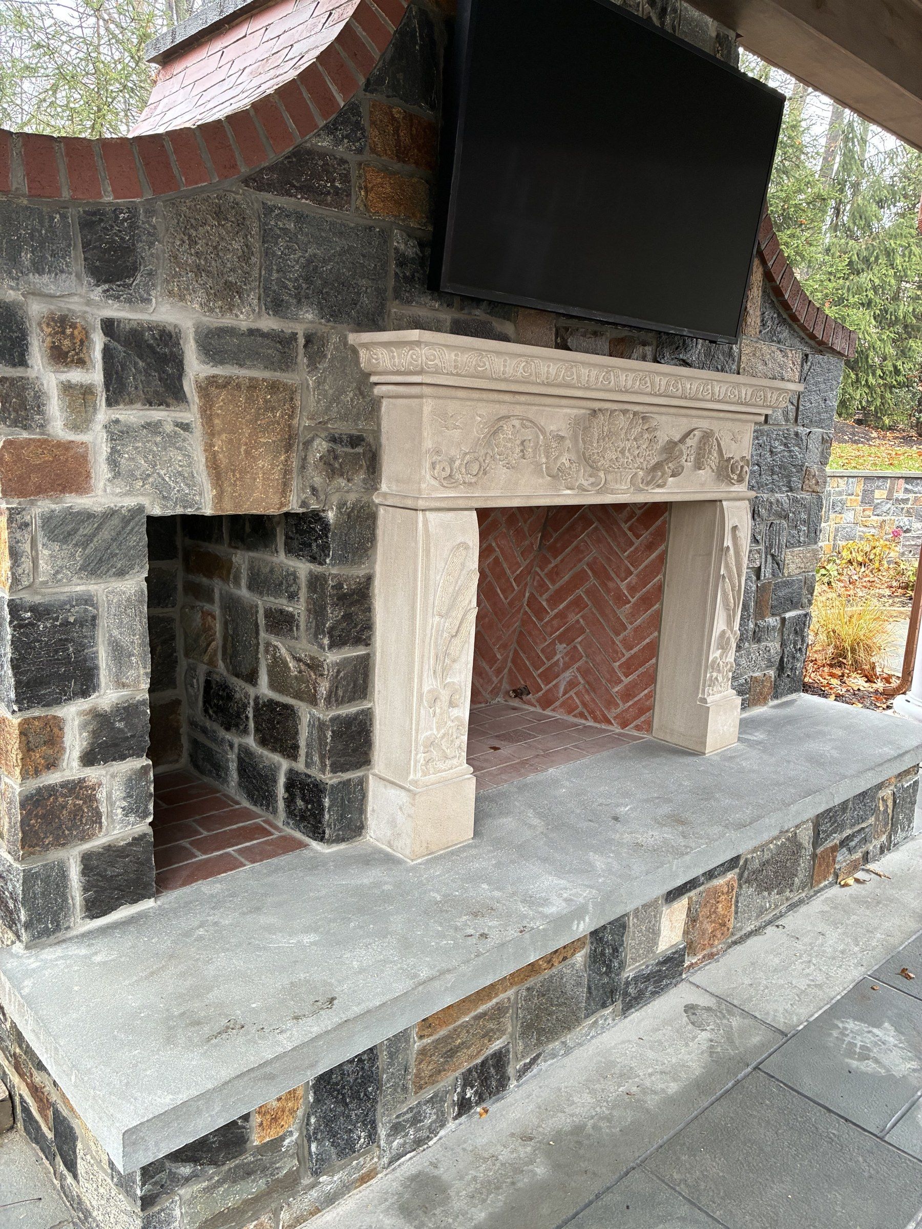 Three-quarter view from the right of the outdoor fireplace, showing the depth of the sculpted mantel and the mixed-colour fieldstone surround.