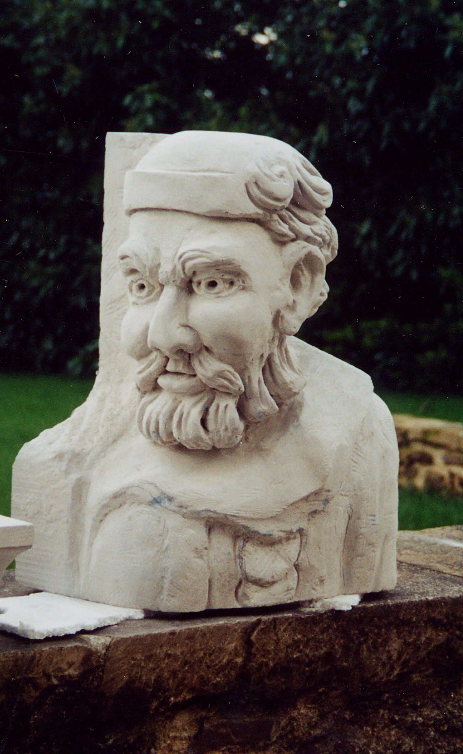 A limestone half-length bust of a bearded man in a round cap, carved in high relief from a block with a flat backing slab, photographed outdoors on a stone wall; the curled beard and furrowed brow are finished, while pencil lines still mark the garment across the chest.