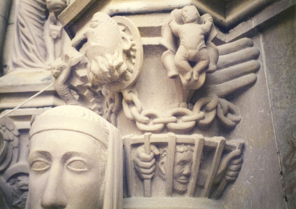 Detail of a carved capital: a bearded head, a small infant figure and a heavy chain carved in the stone, above a band where hands grip the bars of a grille with a face behind it; the head of a statue in the foreground.