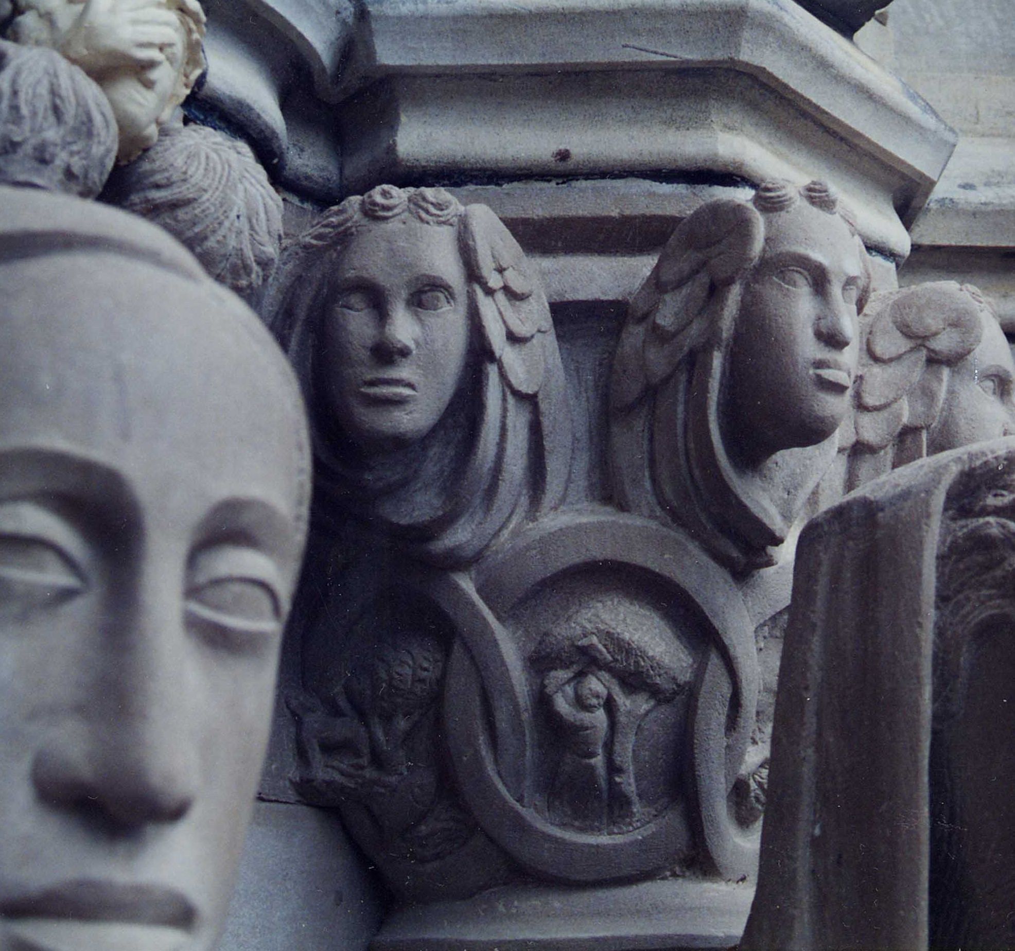 Close-up of a carved capital: young faces with wreathed, wavy hair along the top, above interlaced roundels holding small figures, with the large face of a statue in the foreground.