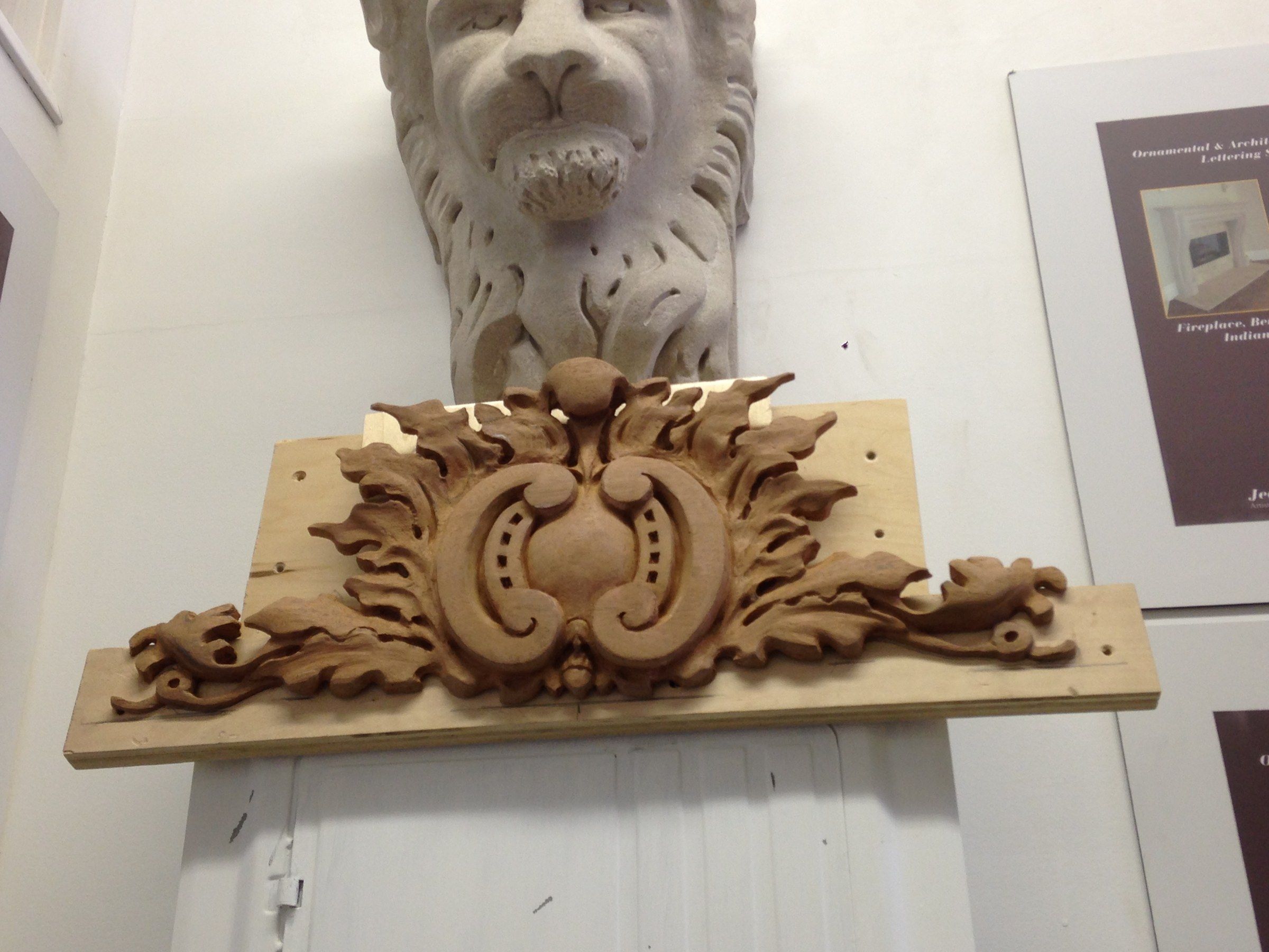 A carved wood cartouche mounted on a white wall beneath a stone lion corbel: an oval center between C-scrolls, framed by curling leaves that spread to each side.