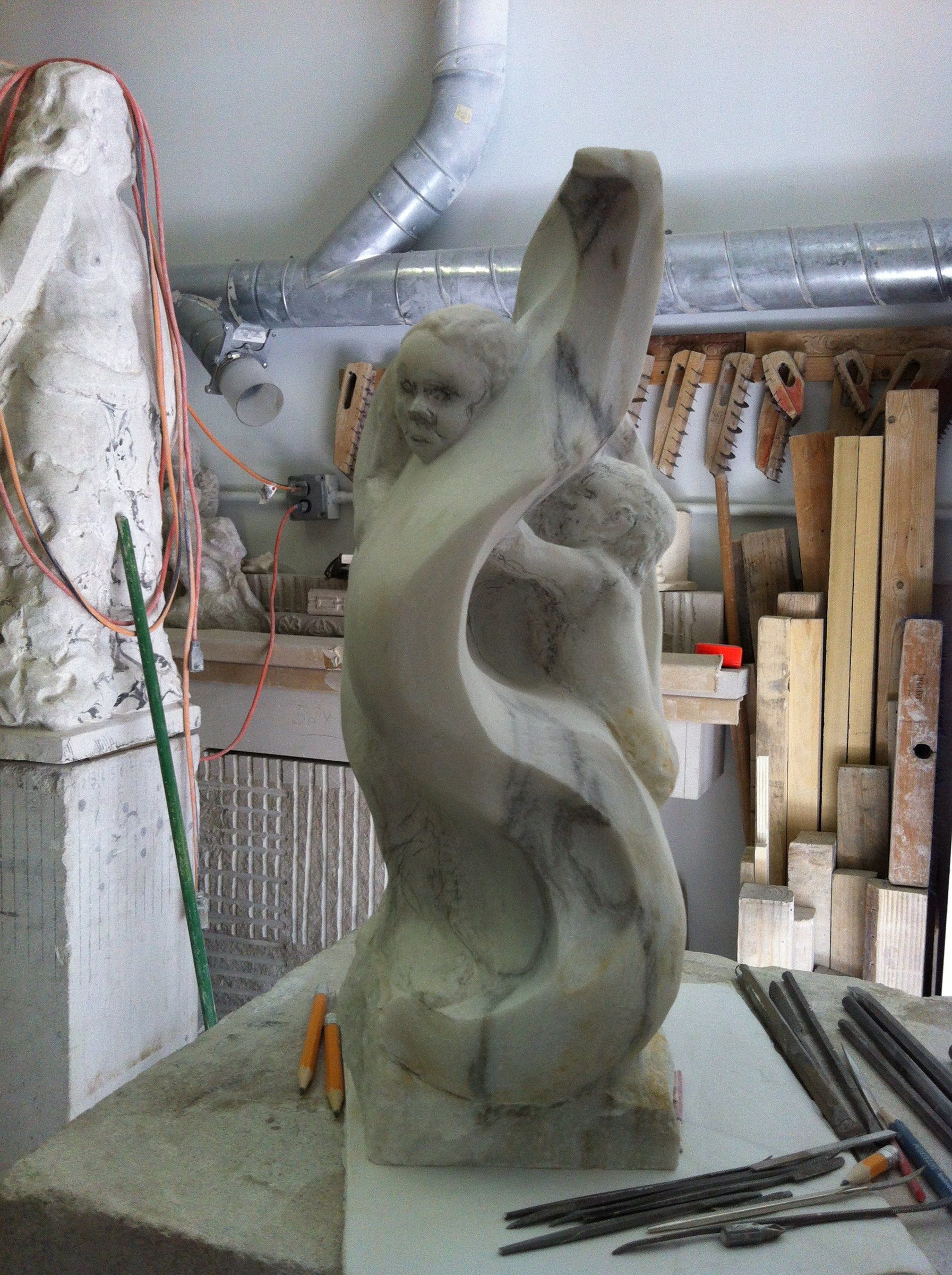 La sculpture élevée en une haute forme torsadée, un visage d'enfant près du sommet et une seconde figure lovée contre elle, des ciseaux sur l'établi.