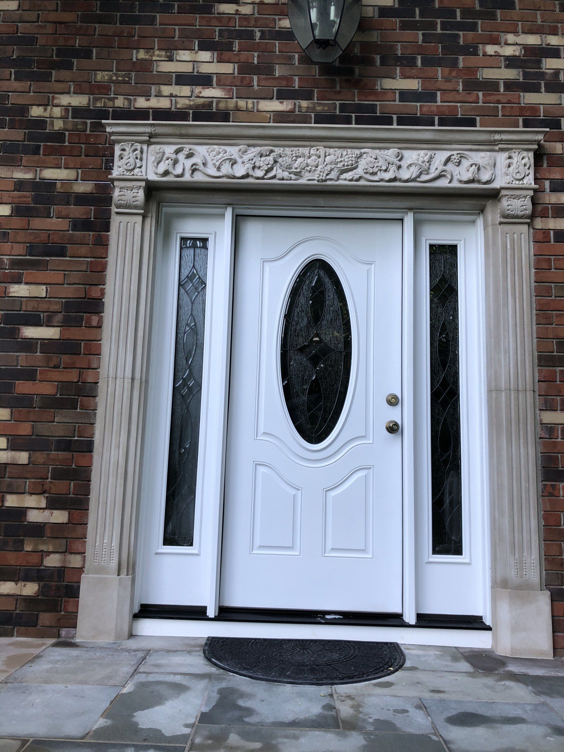 A limestone door surround on a brick house: fluted pilasters on either side of a white front door with an oval glass panel, under a frieze of scrolling foliage and flowers with carved corner blocks.