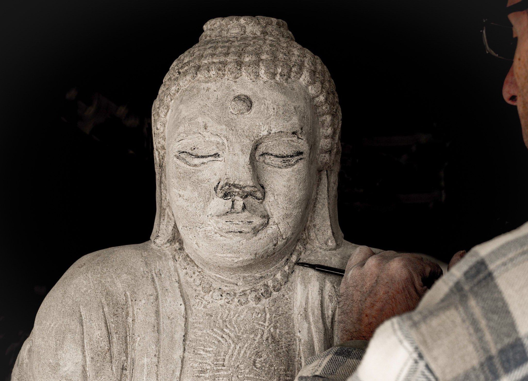 The face of a limestone Buddha with lowered eyelids and curled hair, against a dark background, as a hand in a plaid sleeve works the stone below it with a fine tool.