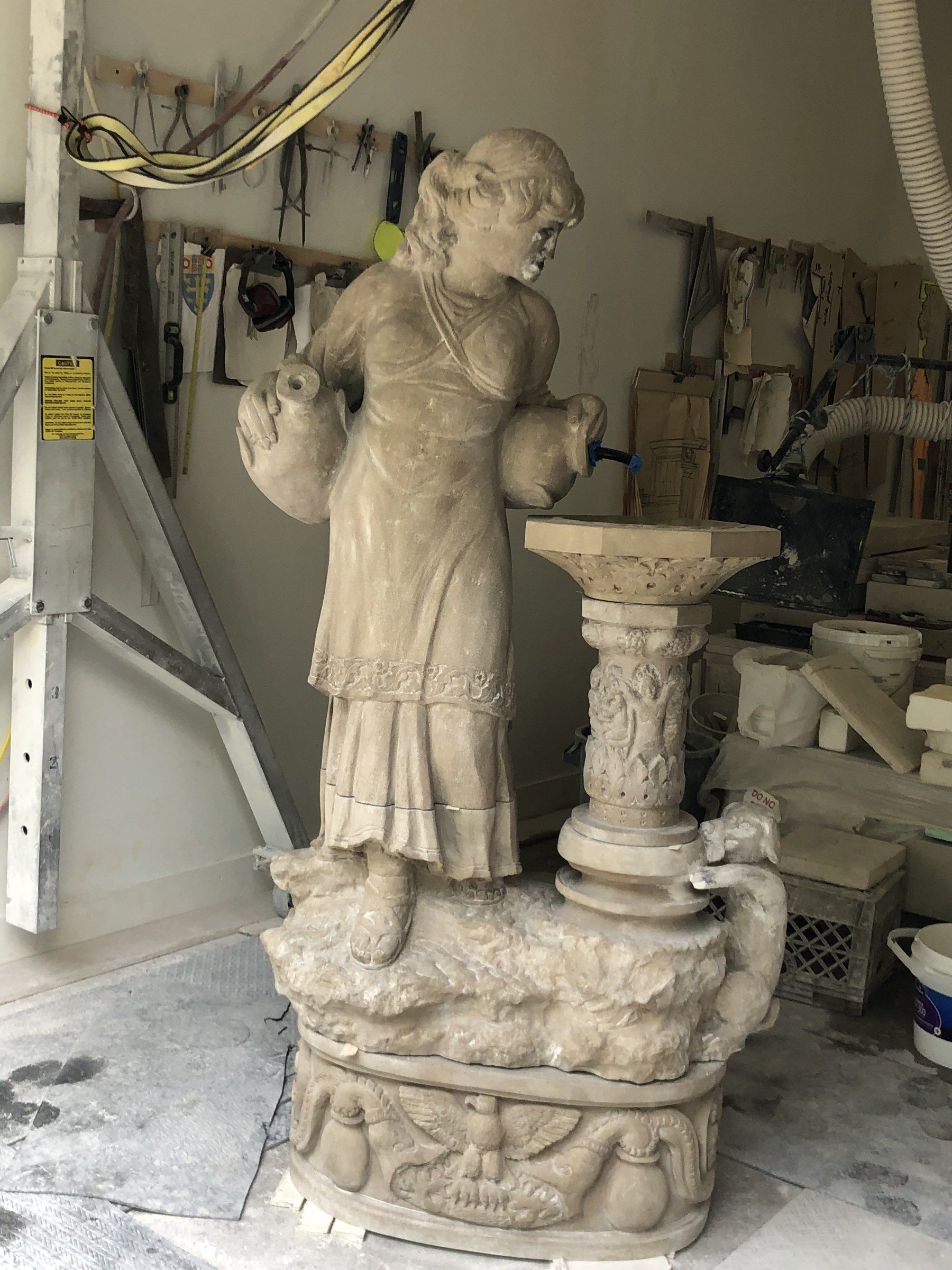 The finished fountain in the studio, seen from the front: the figure, the carved column and basin, a small animal climbing at the foot of the column, and the oval pedestal carved with an eagle and urns.