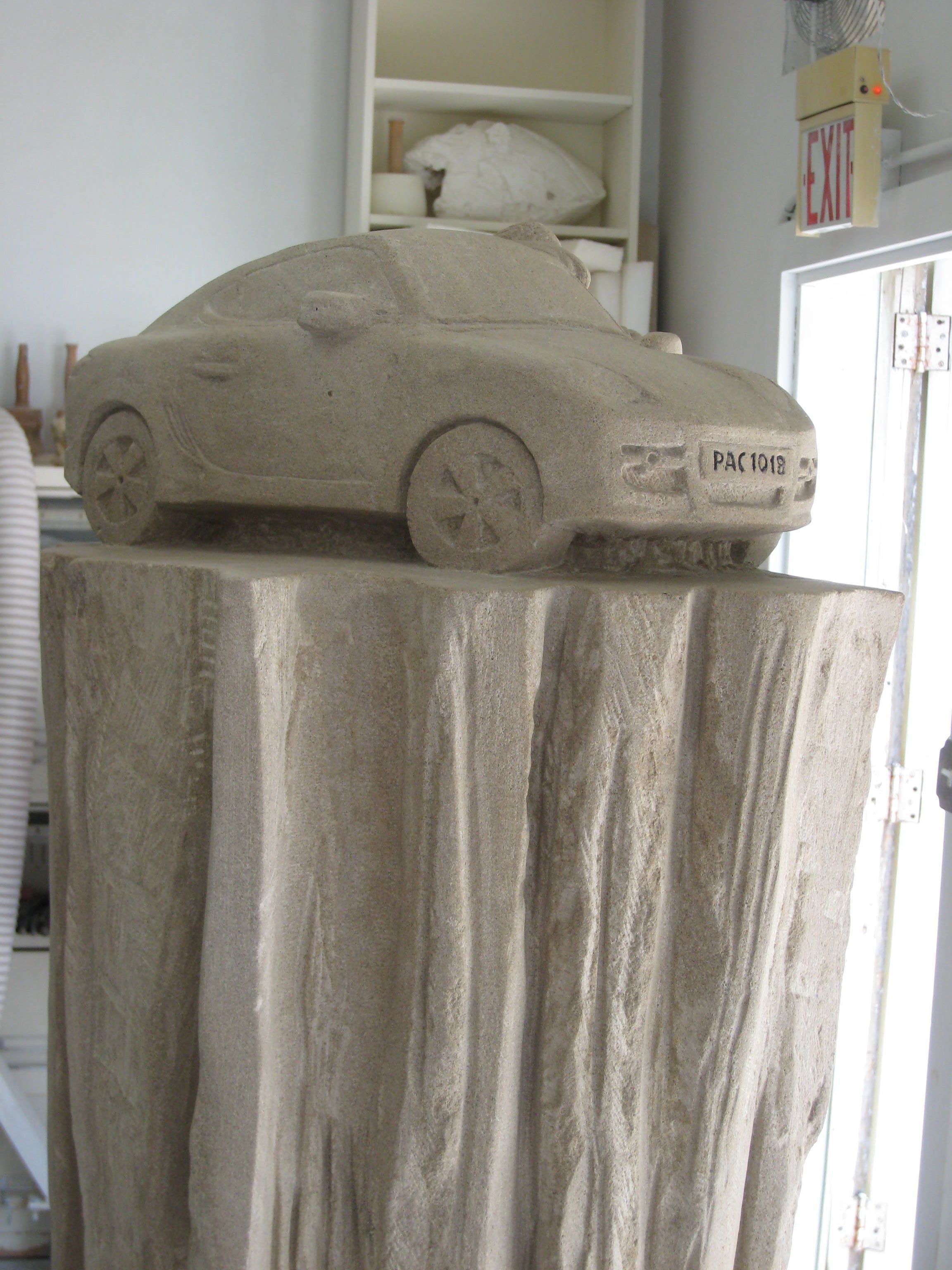 Carved limestone sports car at full scale, with sculpted wheels and a hand-carved licence plate reading PAC 1018, displayed on a draped stone pedestal.