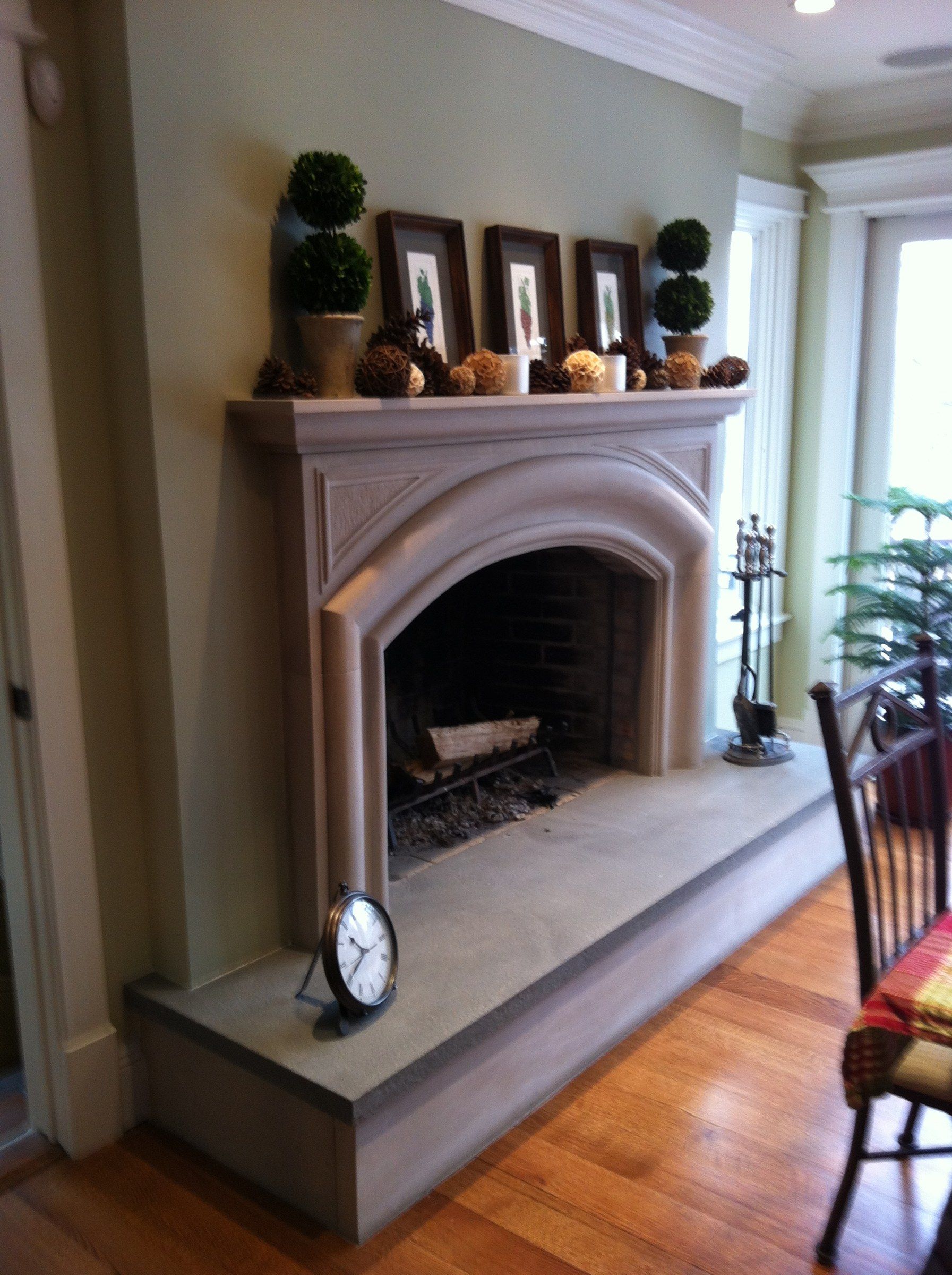 The fireplace seen at an angle, with a clock on the raised hearth and a window to the side.