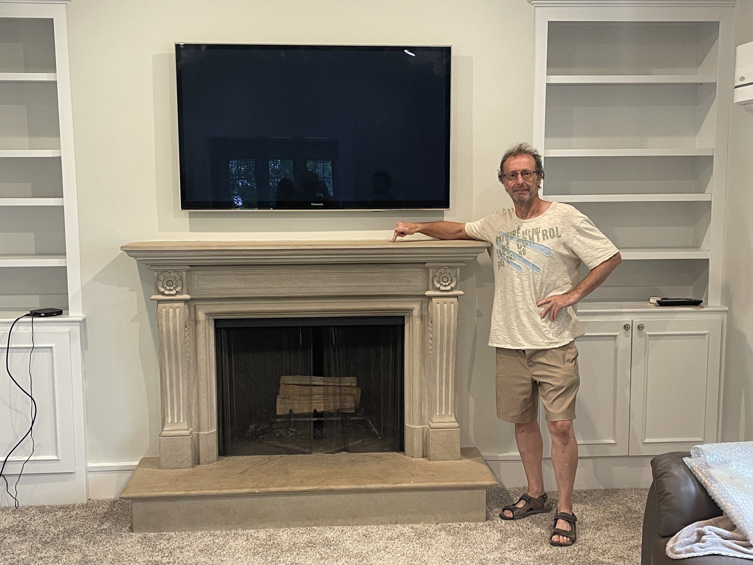 Jean-Claude Marchionni standing beside a limestone fireplace, one hand on the mantel: fluted pilasters topped with rosettes, a raised hearth, built-in shelves on either side and a television above.