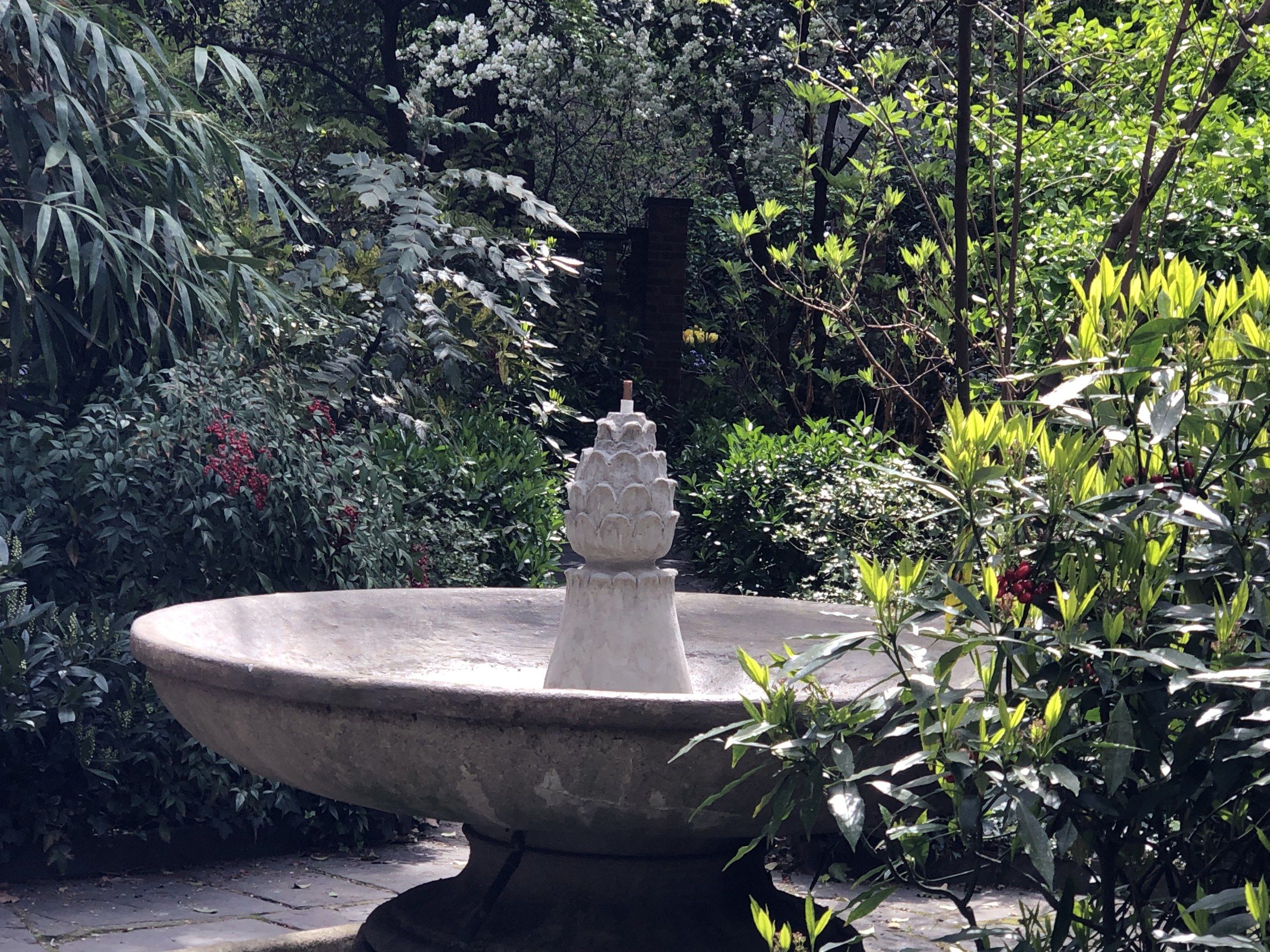 The fountain seen at an angle among the plantings, the finial rising from the center of the basin.