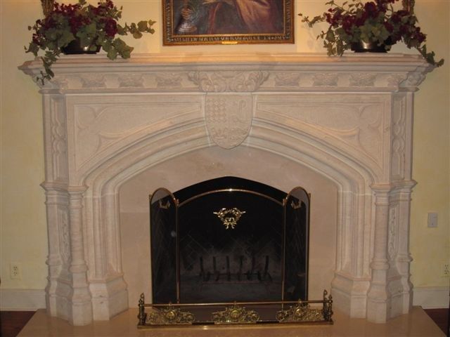 A carved limestone interior fireplace with a trefoil-arched opening framed by gothic pilasters, topped by a mantel bearing a central heraldic coat of arms, with pinnacled uprights at each side and a brass firescreen below.