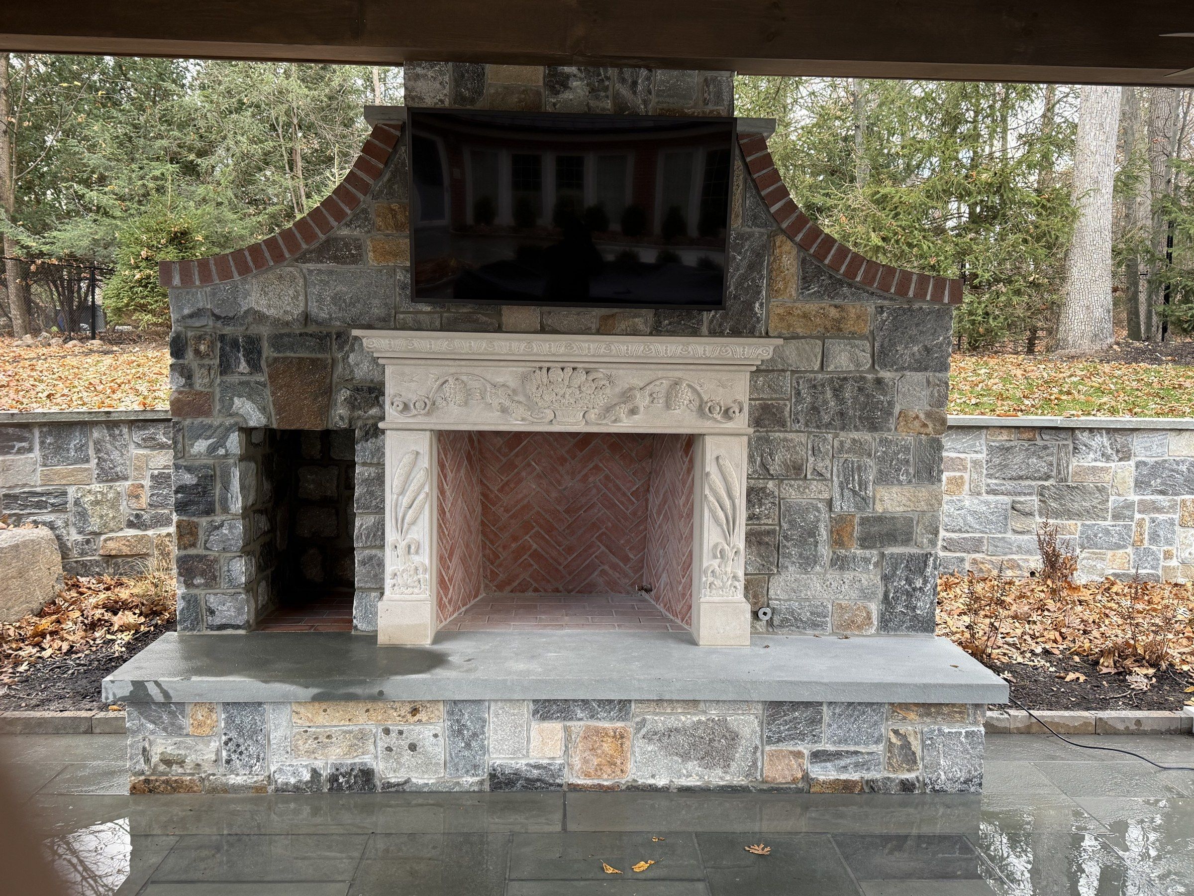 A large outdoor fireplace in a private patio in Bergen County, New Jersey: a carved limestone mantel in cream, with ornamented pilasters and a sculpted frieze, set within a surround of mixed grey fieldstone, with a flat-screen television mounted above.