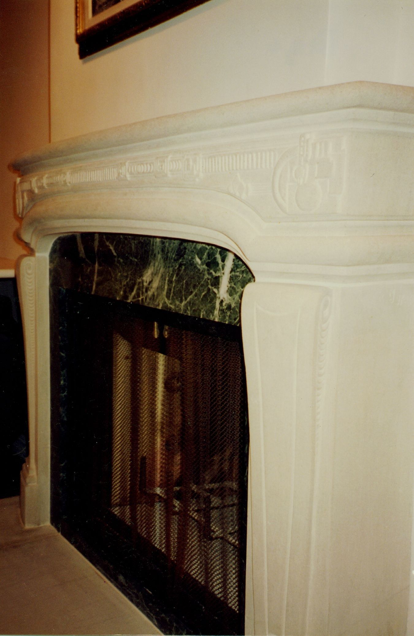 A pale limestone fireplace seen at an angle: a rounded shelf over a curved frieze carved with fluted and geometric panels, a stylized geometric motif in each corner, tapered jambs with recessed panels and beaded scrolls, and a dark green veined marble slip framing a mesh fire screen.