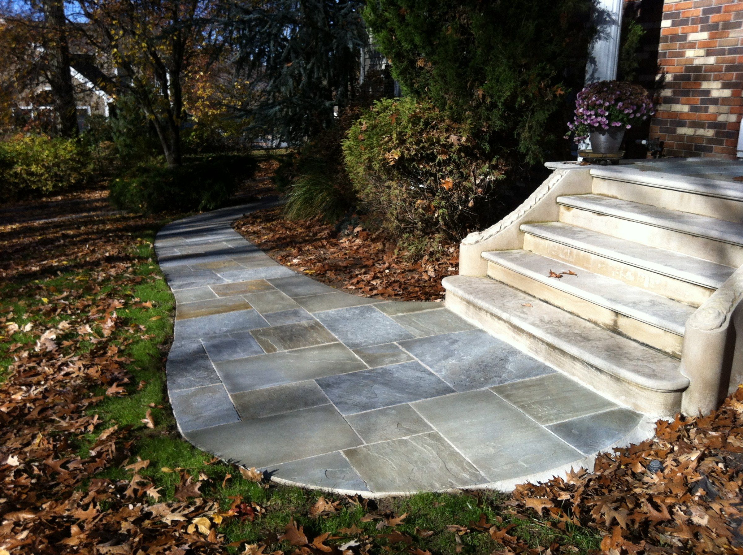 Limestone garden steps with rounded treads and scrolled side walls, leading up to a brick house from a curving path of stone paving among fallen autumn leaves.