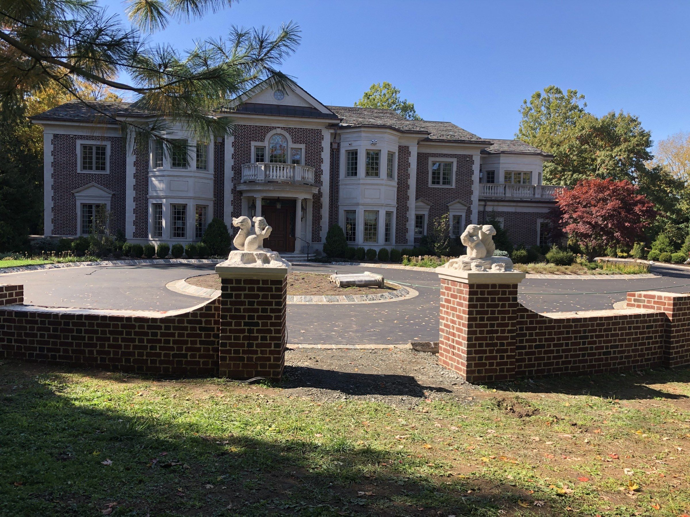 The two squirrels in place on brick gate piers at the entrance of a circular driveway, in front of a large brick house with white trim, under a blue sky.