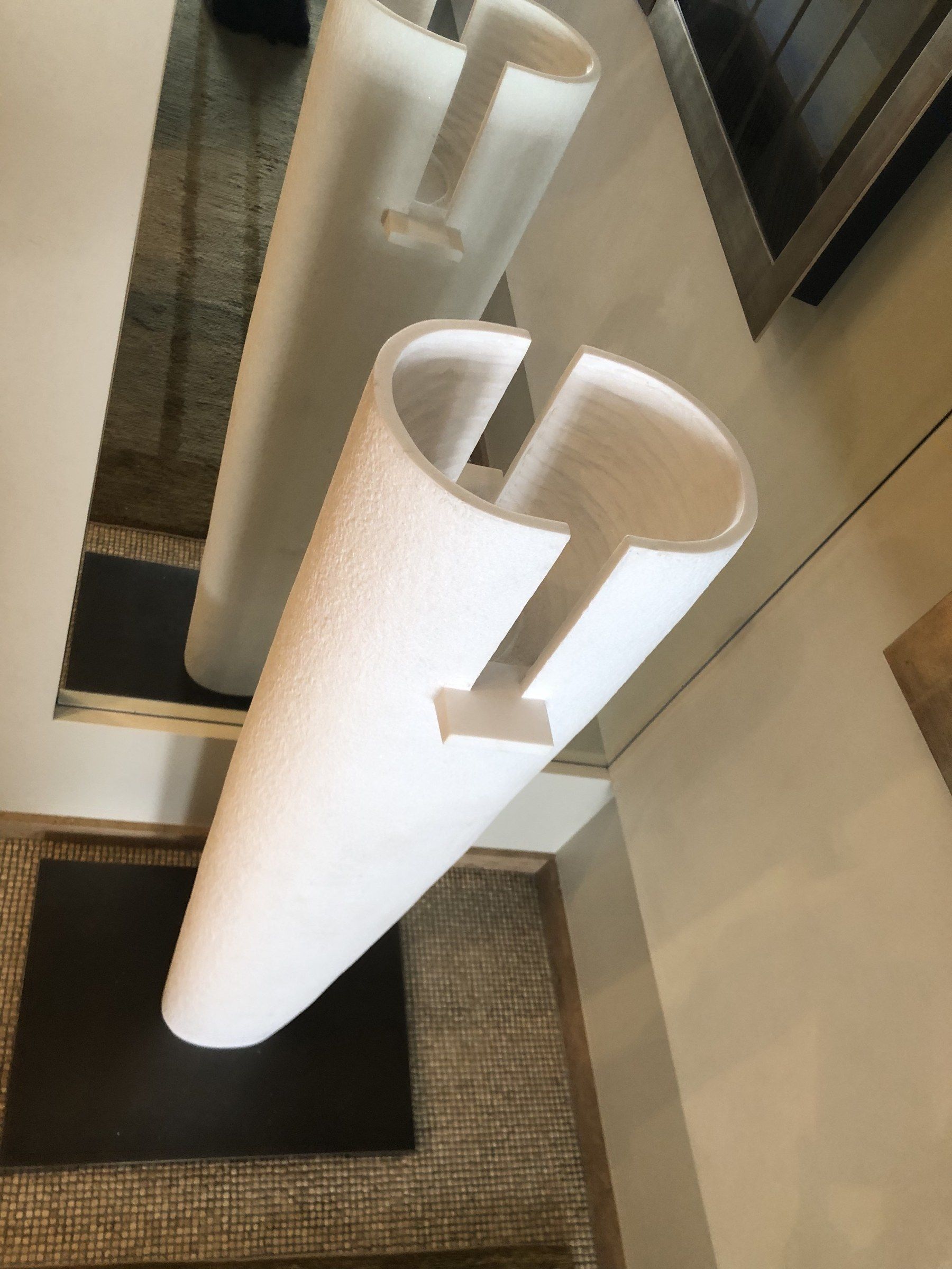 La sculpture installée en avril 2025, vue en plongée, de biais : le cylindre creux à paroi fine, ouvert au sommet, une fente descendant depuis le bord, et son reflet dans le miroir derrière.