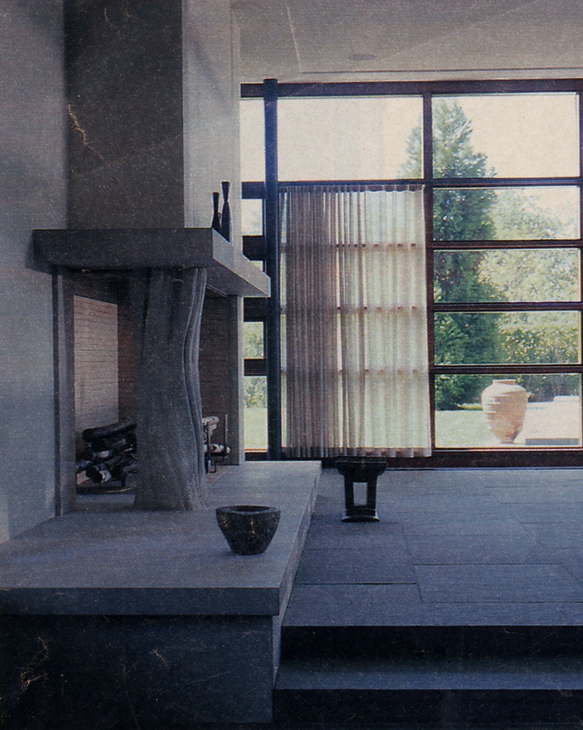 Hand-carved stone fireplace shaped as a weathered tree trunk, rising from a monolithic stone hearth in a minimalist East End interior with full-height garden windows.