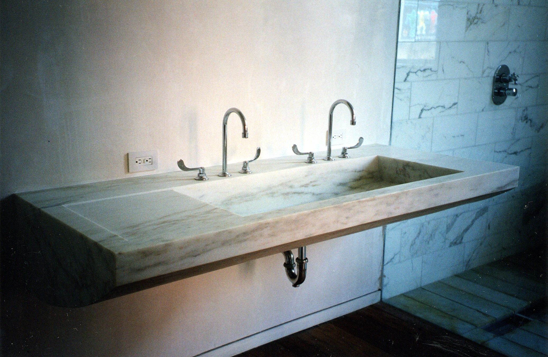 A wall-hung sink cut from a single slab of white marble with soft grey and amber veining: a long trough basin sloping toward the drain, a shallow recessed panel at the left end, two gooseneck faucets along the back edge, and a rounded underside where the slab meets the wall.