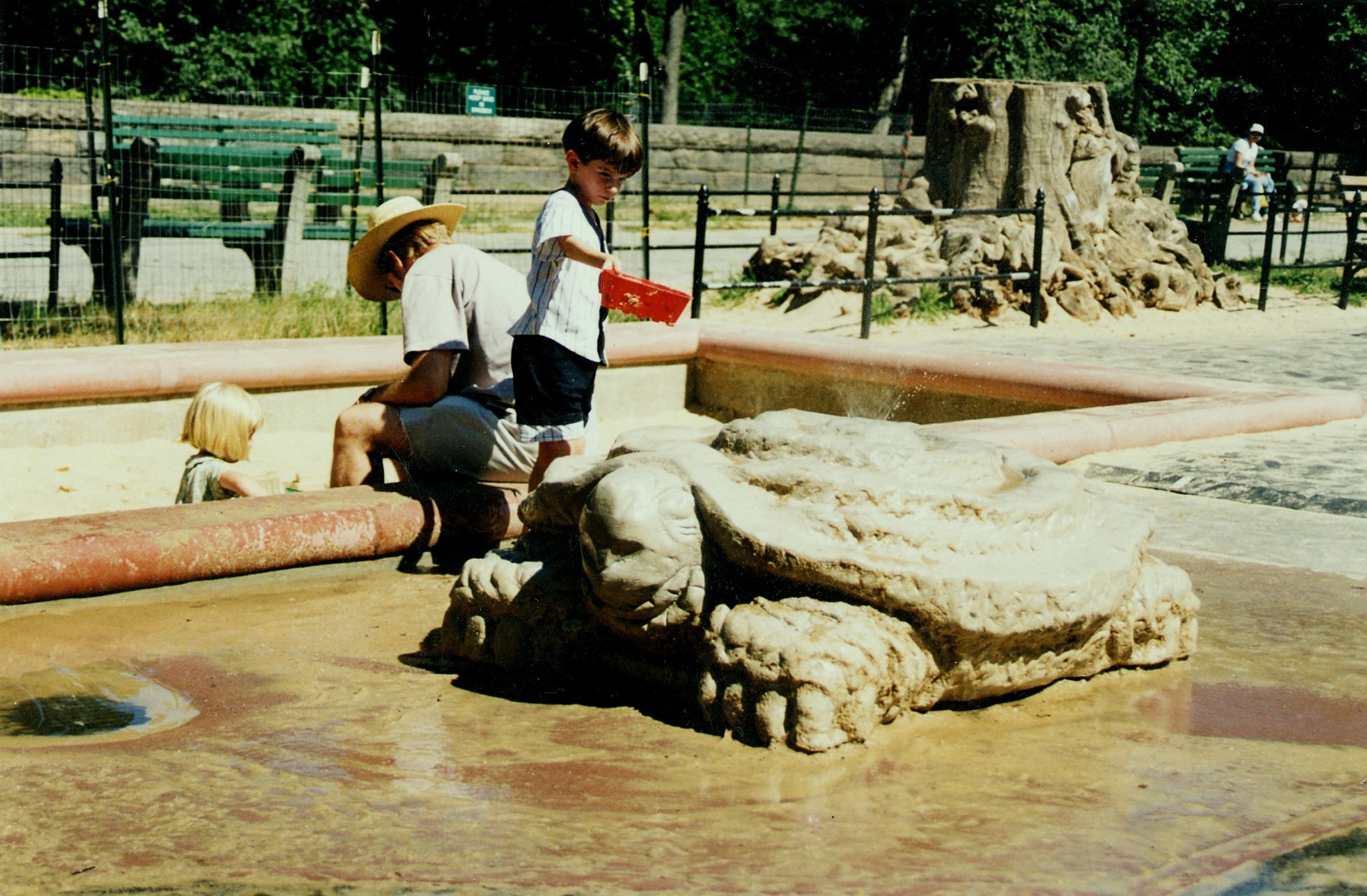 A large carved limestone turtle set in the shallow splash pool of a city playground, a jet of water rising from its stepped, spiraling shell; children play at the pool's edge, with green park benches, a black iron fence, and a rough stone wall behind.