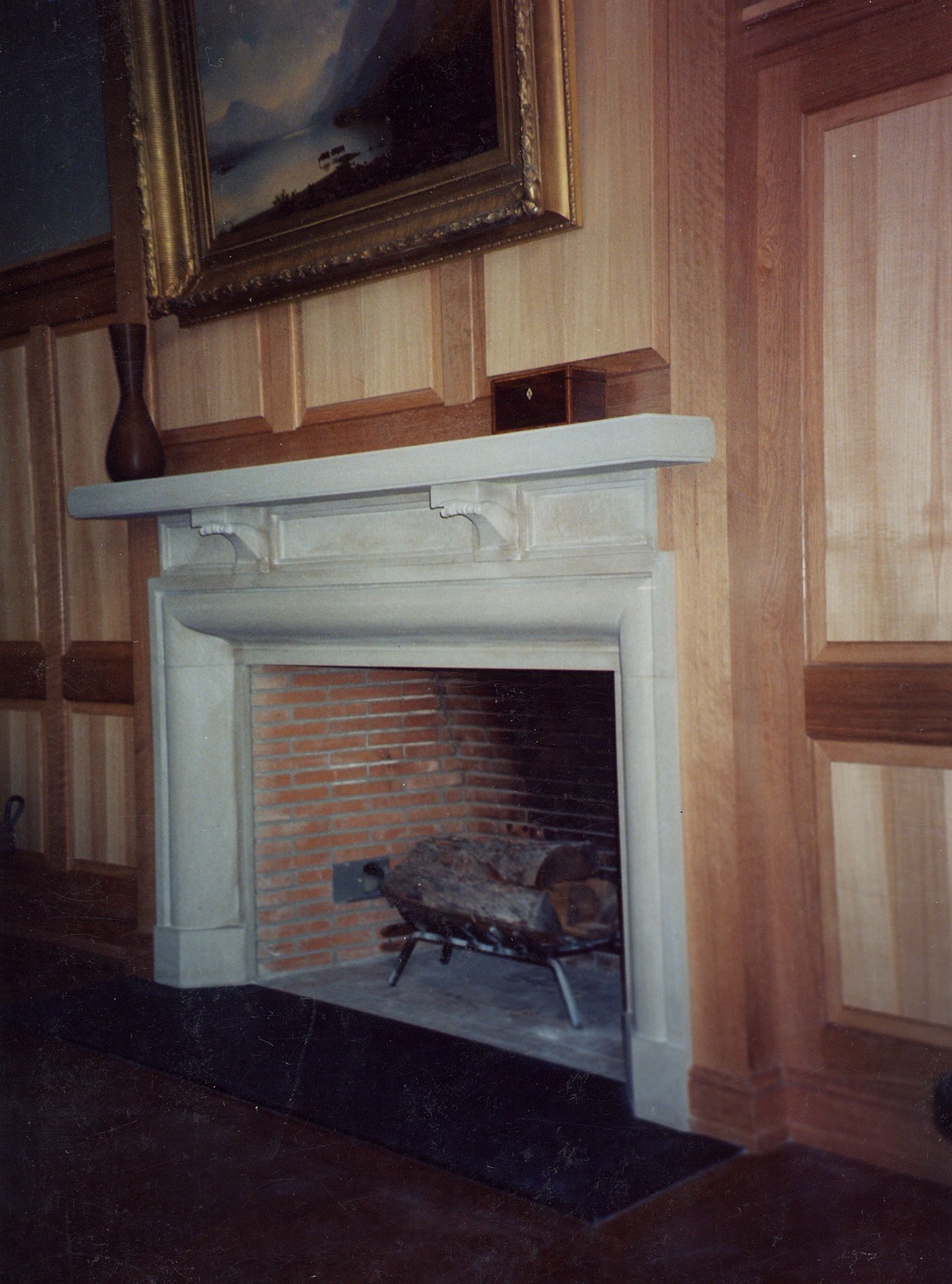 A pale limestone fireplace set into oak-paneled walls: a broad bolection-molded surround around a brick firebox with a log grate, beneath a plain mantel shelf carried on two small carved corbels, with a dark stone hearth at its base.
