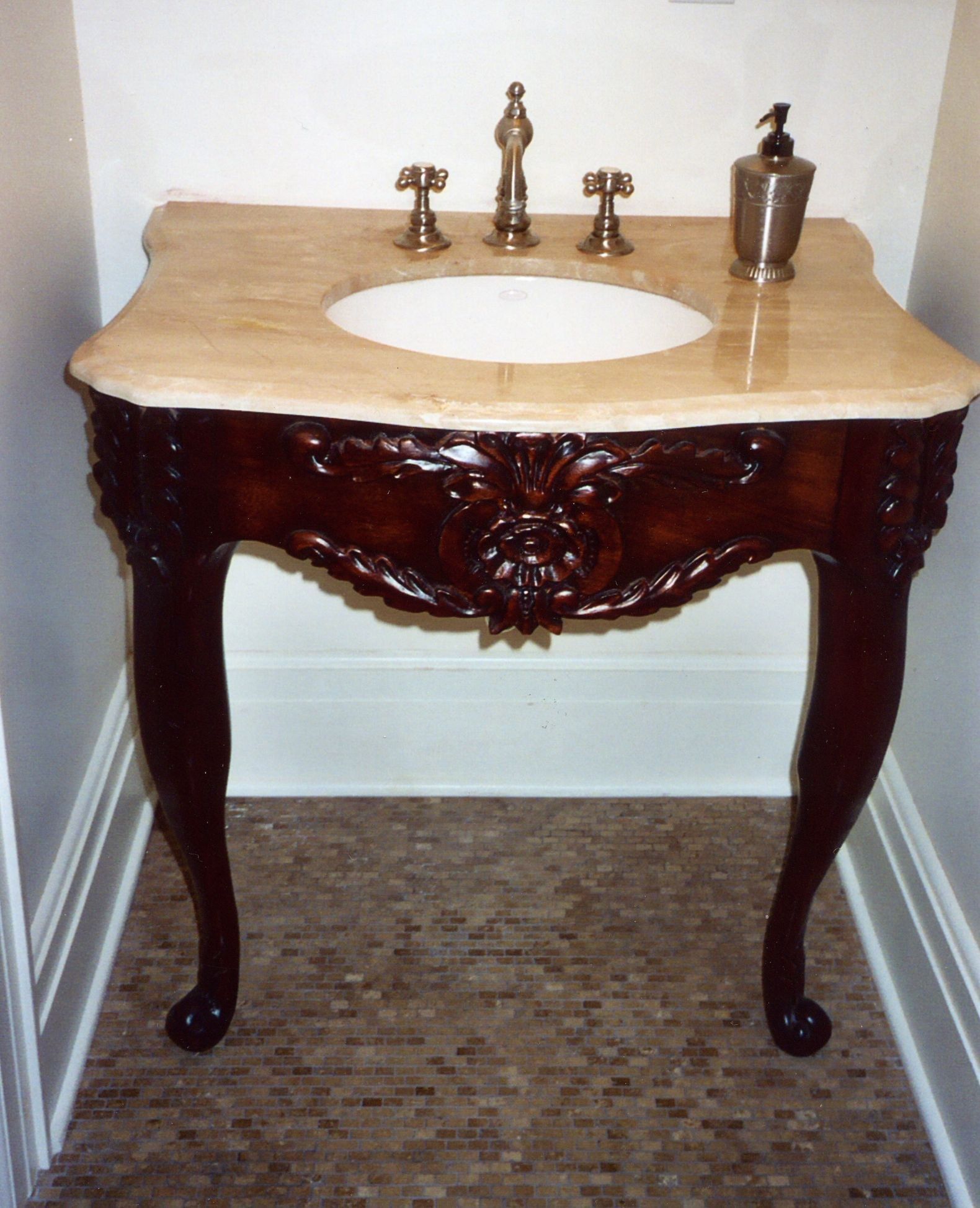 A carved mahogany vanity in a small powder room: a dark, polished base on slender cabriole legs, its serpentine apron carved at the center with a full rosette between sprays of acanthus and leafy garlands, and acanthus carved down the corner blocks; above, a shaped cream marble top with an undermounted white basin and cross-handle faucets.