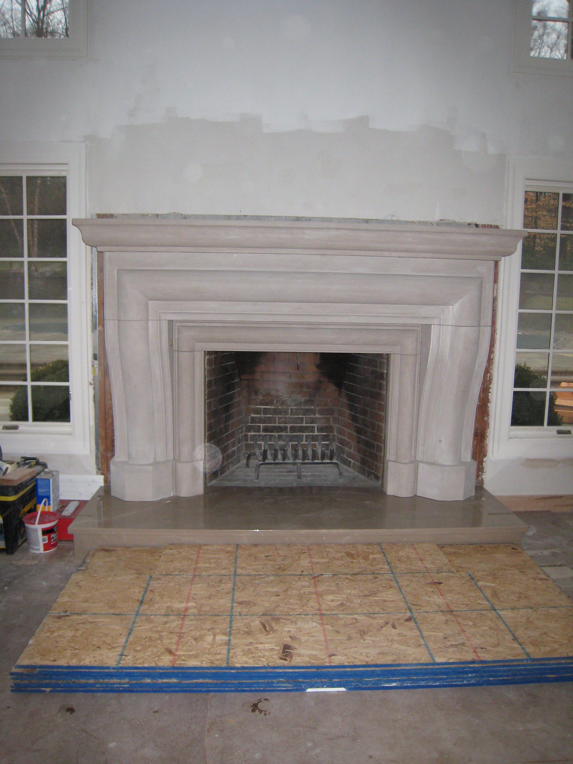Carved limestone fireplace with mouldings and a scroll-shaped mantel, installed in a residential interior with tall windows on either side.