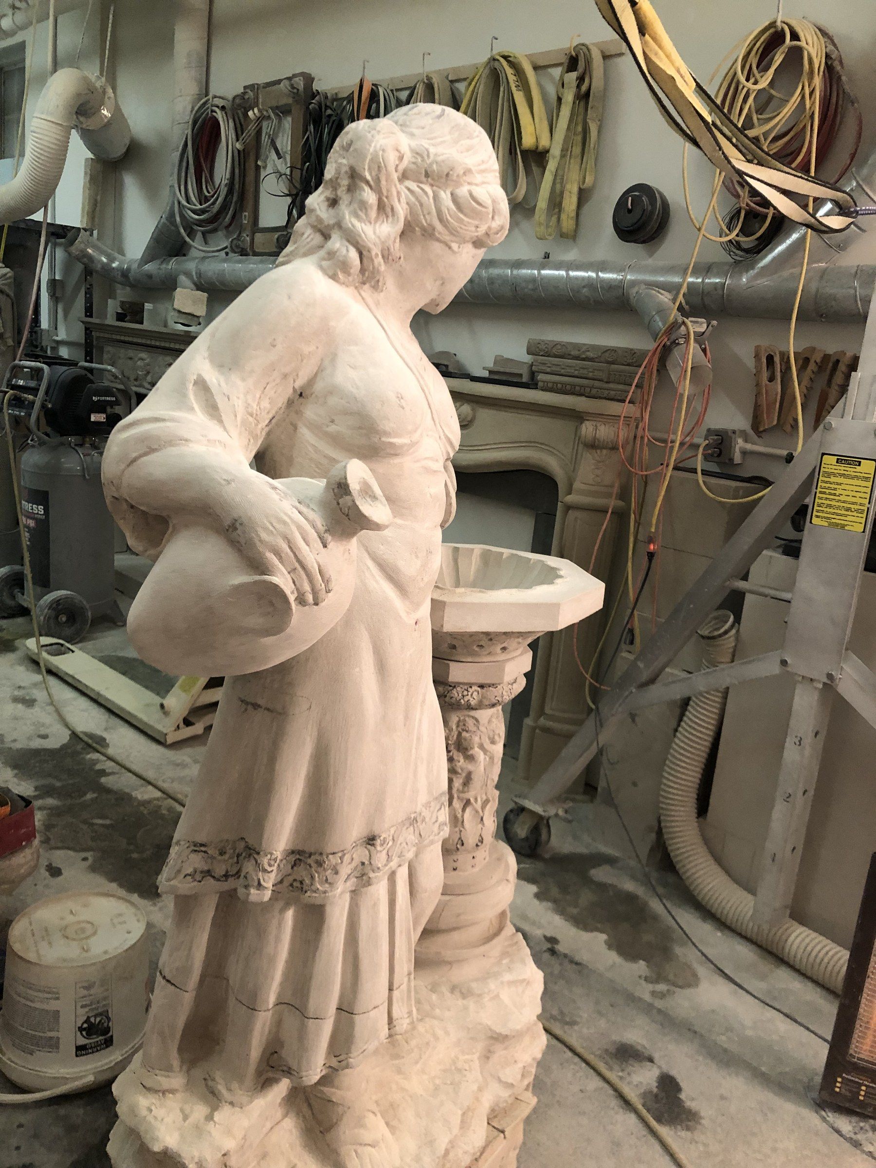 Close view of the figure from the side, a jug held against her hip, her dress finished with a carved lace border.