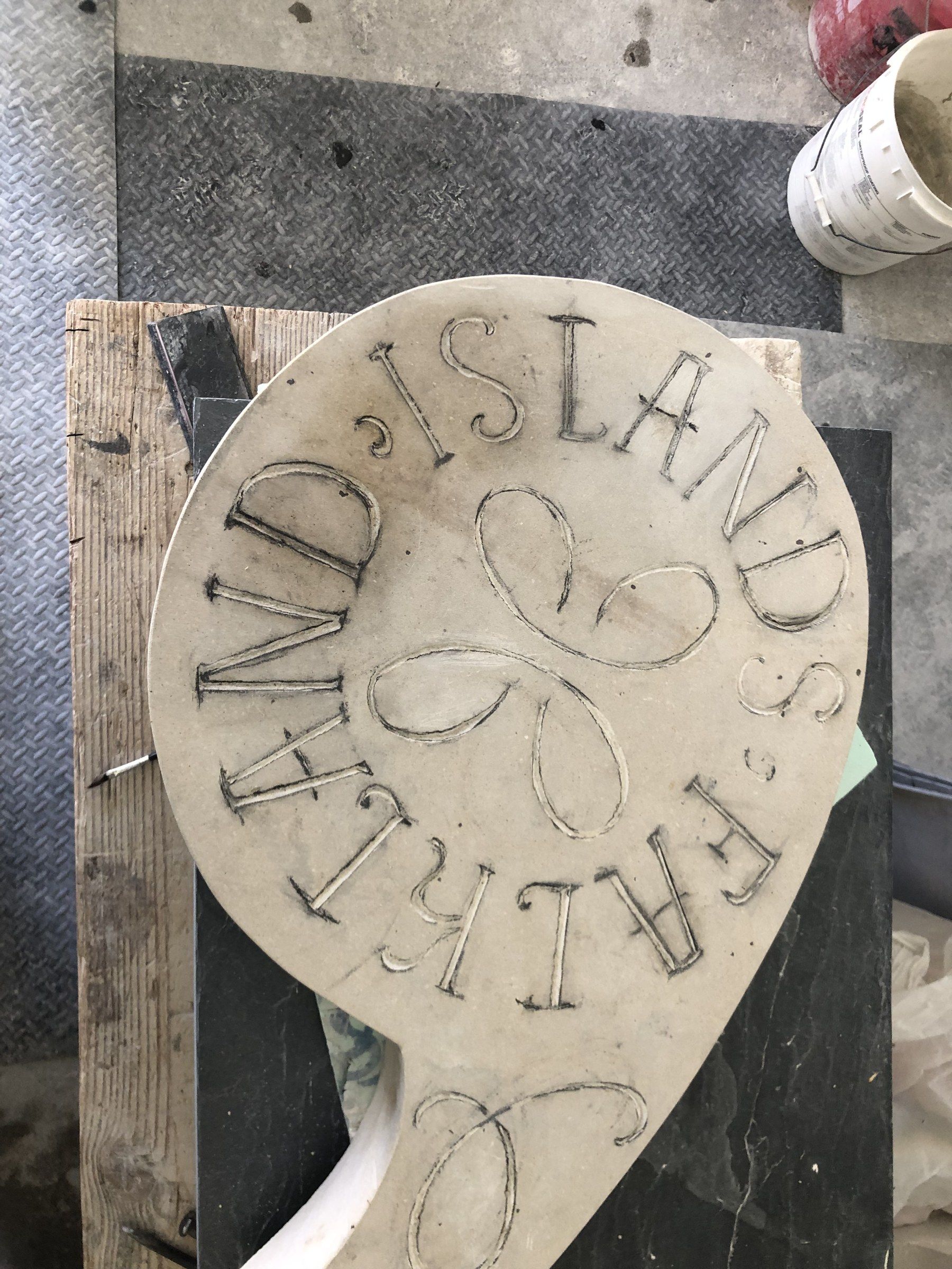 The plaque on the workbench with its lettering and loops outlined in pencil, ready to be cut.