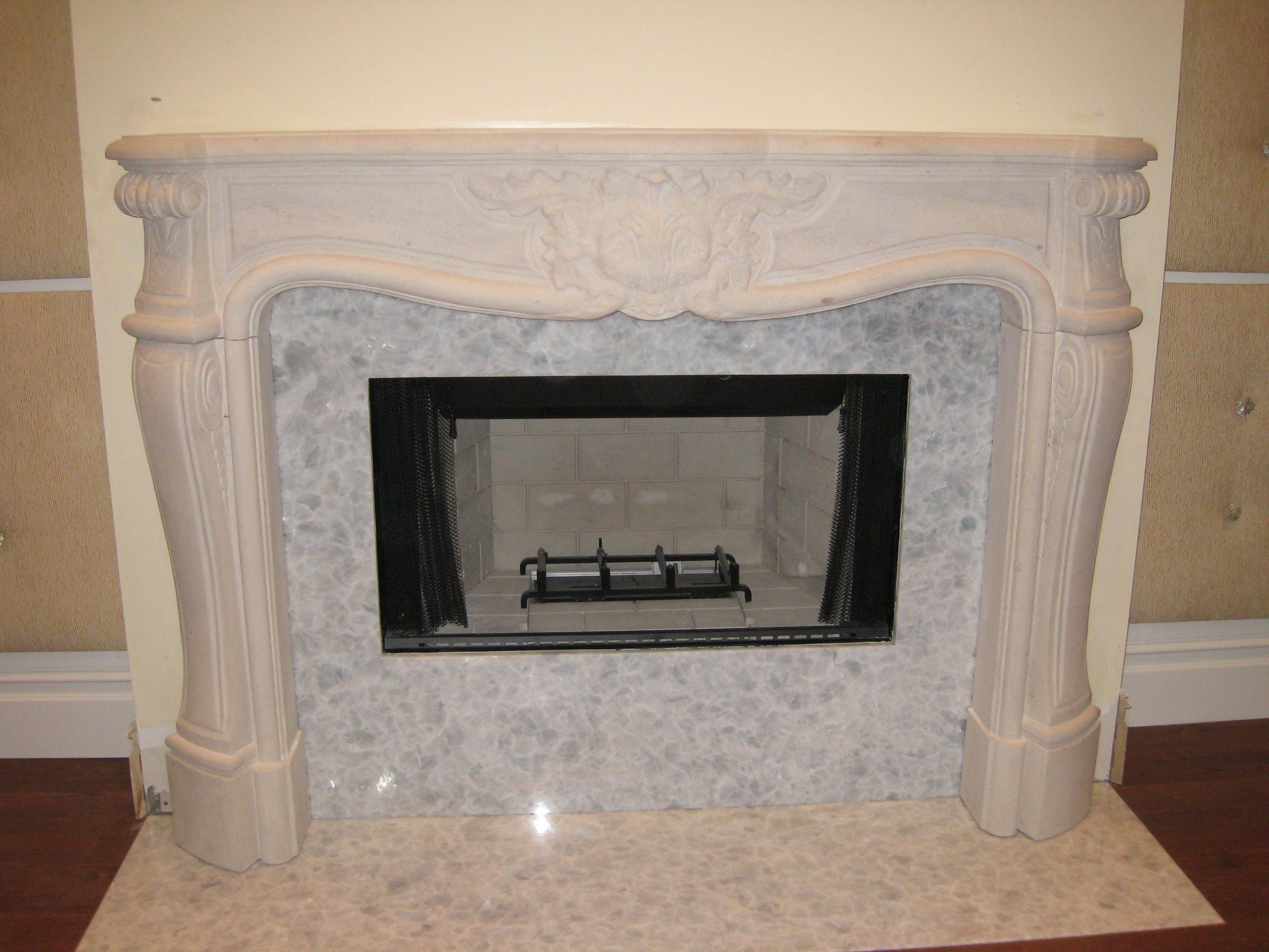 A curved limestone mantel with a carved shell-and-leaf centerpiece, around a firebox framed in pale pebble tile.