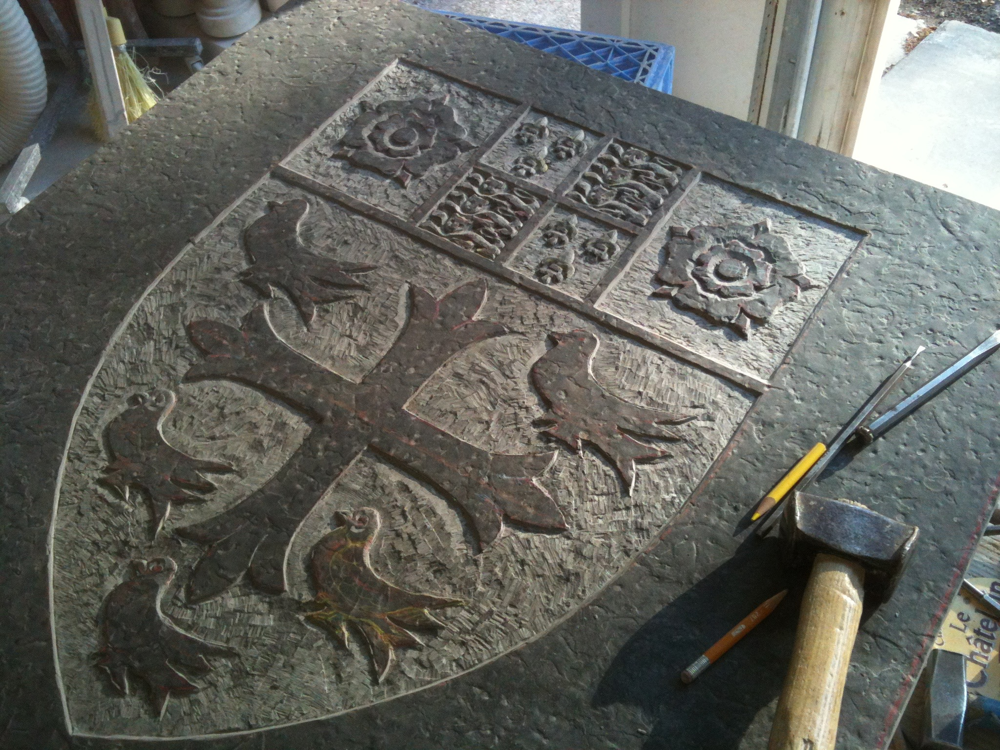 Carved Welsh slate heraldic shield with doves, a central cross, and tracery panels, resting on a workbench beside a mallet and chisels.