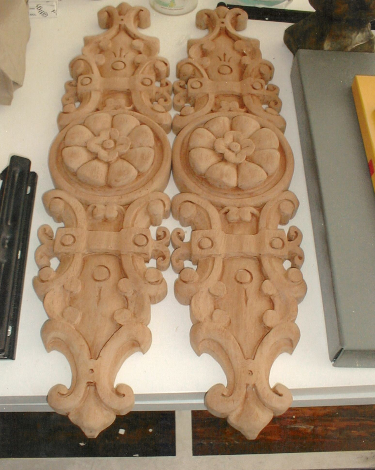 A matching pair of long, unfinished carved wood appliqués laid side by side on a workbench: each rises from a pointed pendant through a scrolled cartouche and a strapwork band studded with round bosses to a central roundel holding a rosette, then a second strapwork band and a scrolled crest.