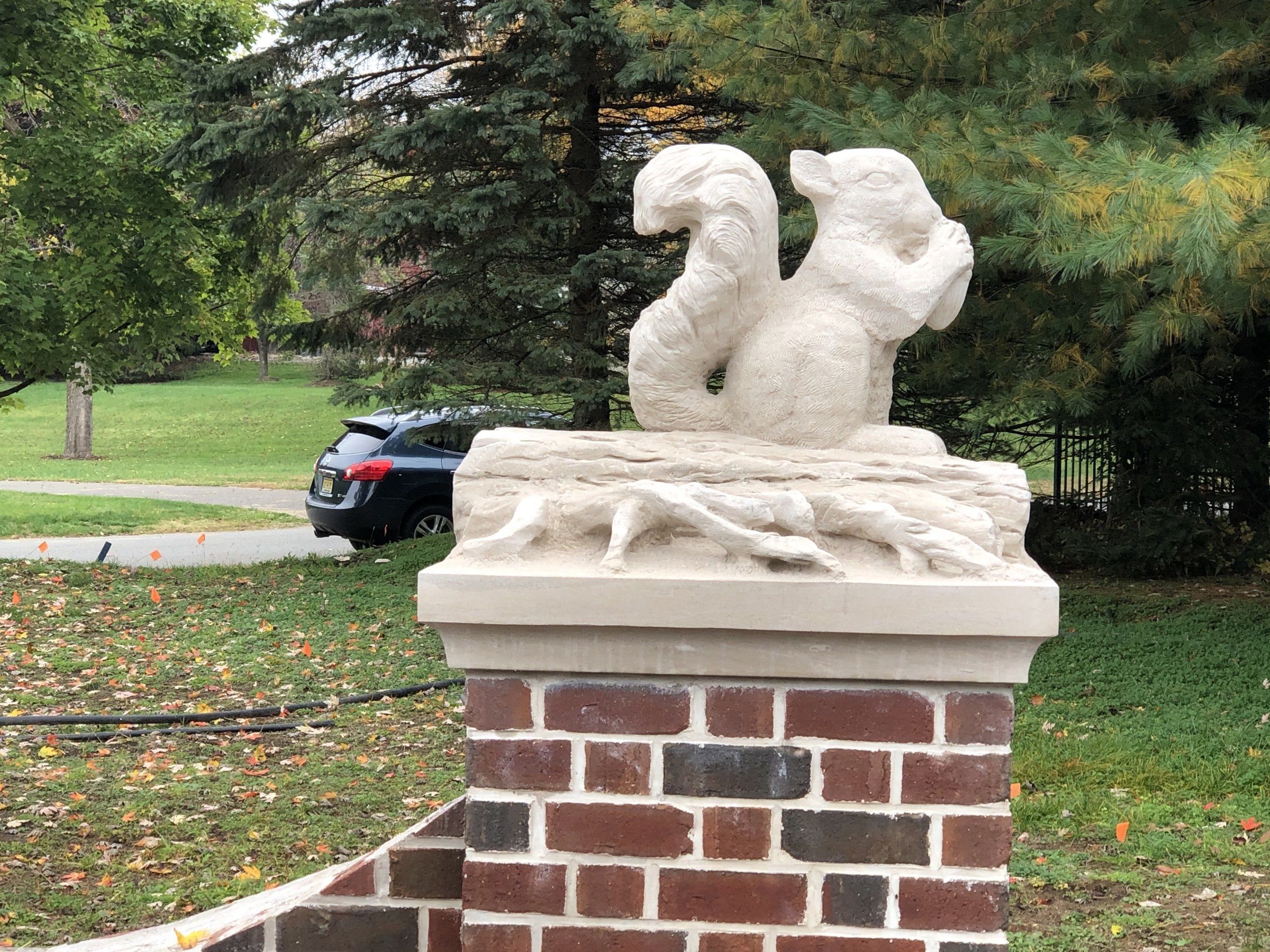 The squirrel on the log, seen from the front on its brick pier, with evergreens and a lawn behind.