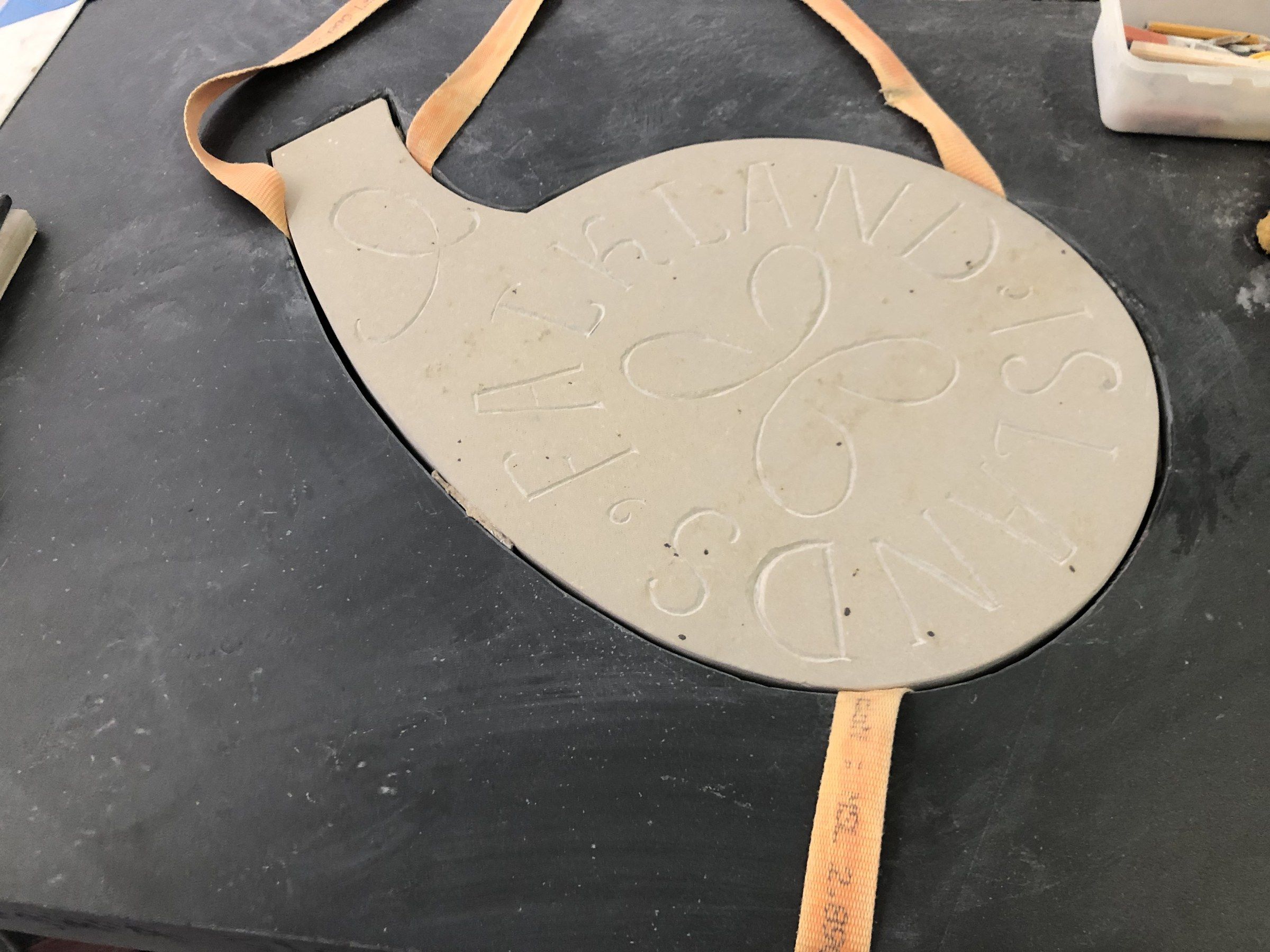 A sandstone plaque shaped like a rounded teardrop, lettered “FALKLAND ISLANDS” around a central motif of interlaced loops, set into a dark stone slab with lifting straps still around it.