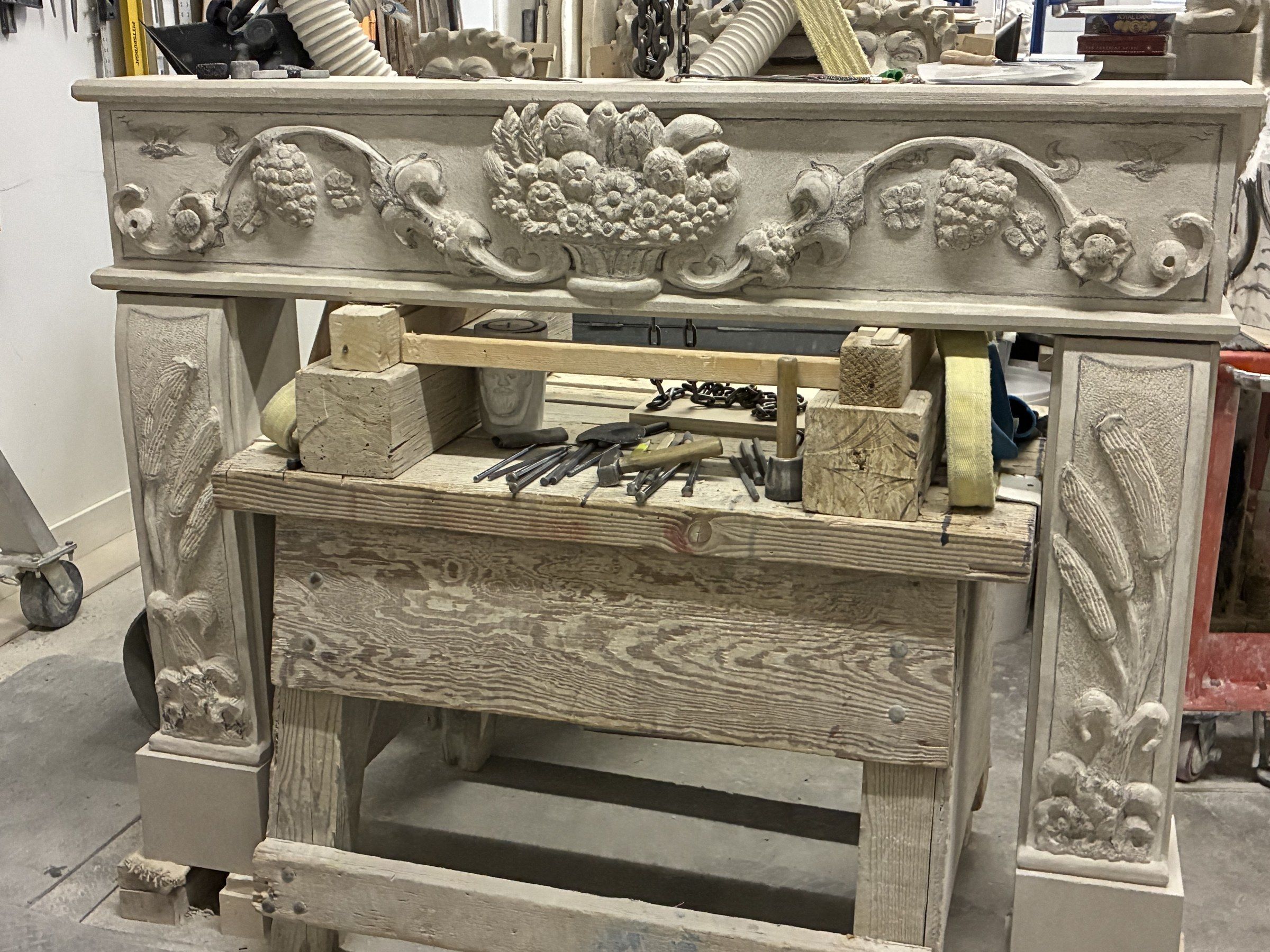 July 2025: the mantel assembled on trestles in the studio, the frieze carved with a central basket of fruit, grape clusters, scrolling vines and rosettes, between jambs carved with ears of grain.