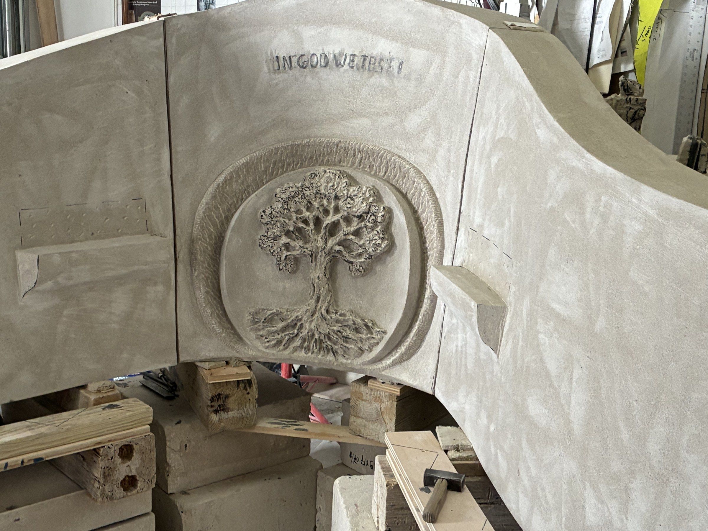 The inscription “IN GOD WE TRUST” carved at the summit of the central panel in August 2026, above the Tree of Life medallion.