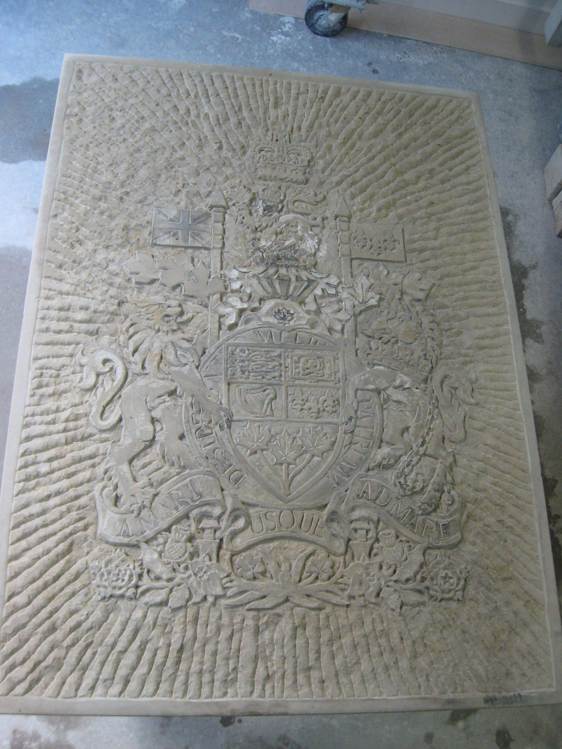 A rectangular sandstone relief of the arms of Canada lying in the studio: lion and unicorn supporters, a shield, a crown and flags, on a background of radiating chisel strokes, above a ribbon reading “A MARI USQUE AD MARE.”