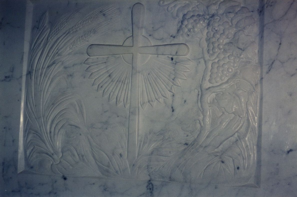 The carved front panel of a white veined marble altar: a plain cross set against a radiating sunburst at the center, a sheaf of wheat rising along the left edge, and a grapevine heavy with clusters of grapes along the right, all in low relief within a beveled frame.