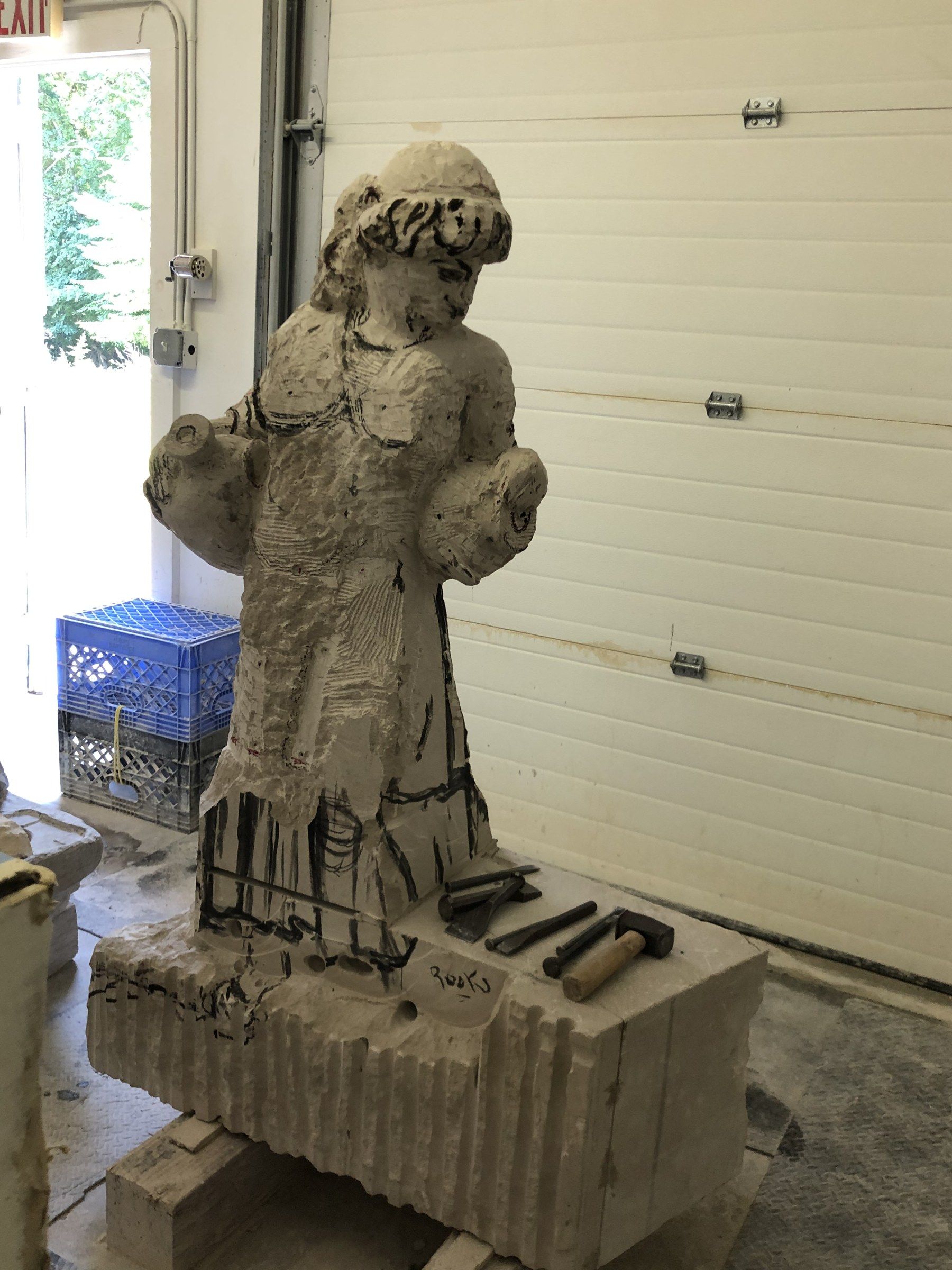 The figure roughed out of the block, covered in guide lines, with mallets and chisels resting on the uncut stone at her feet.