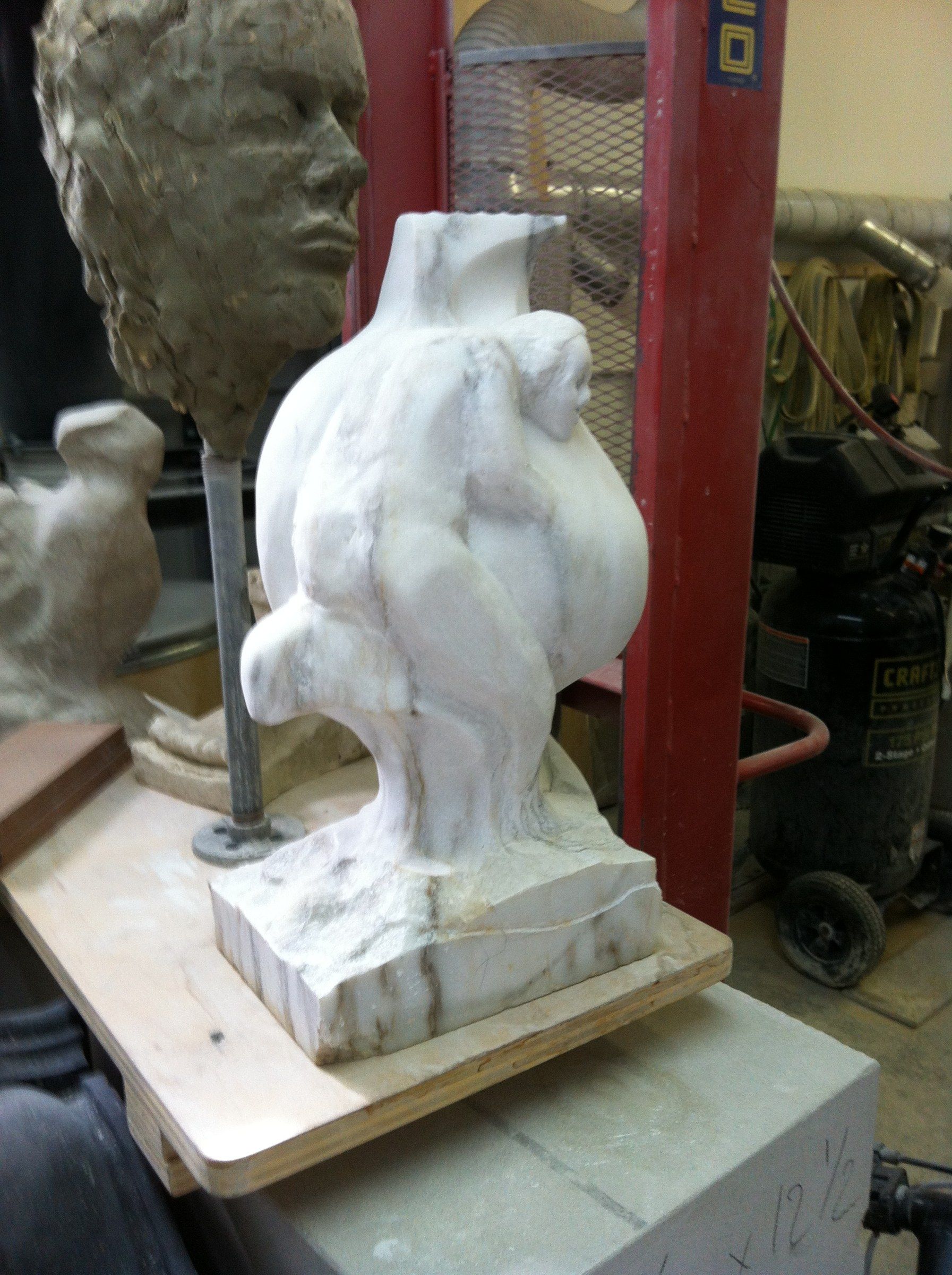 Le marbre à un stade précoce sur une planche de l'atelier : une masse arrondie où une figure commence à apparaître sur le flanc, à côté d'une tête en terre sur une tige.