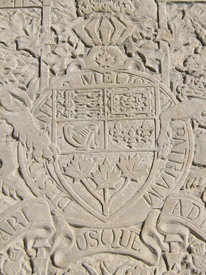 Gros plan de l'écu — trois lions, un lion rampant, une harpe, des fleurs de lys et trois feuilles d'érable — dans un anneau portant « DESIDERANTES MELIOREM PATRIAM ».