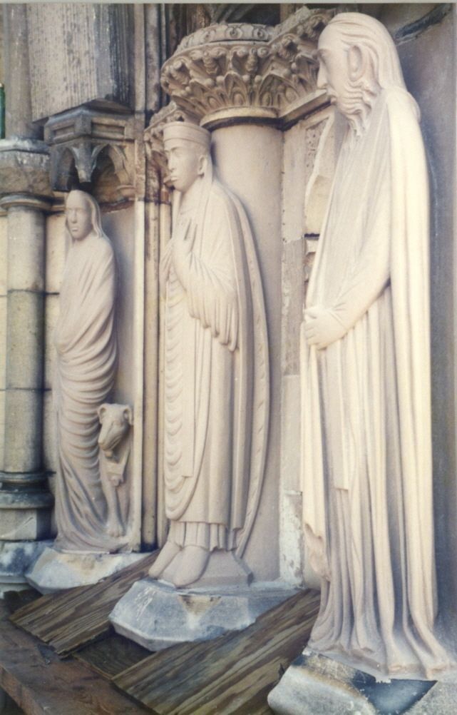 Three tall limestone statues standing on wooden planks in the portal, the new stone still pale, beneath carved capitals.