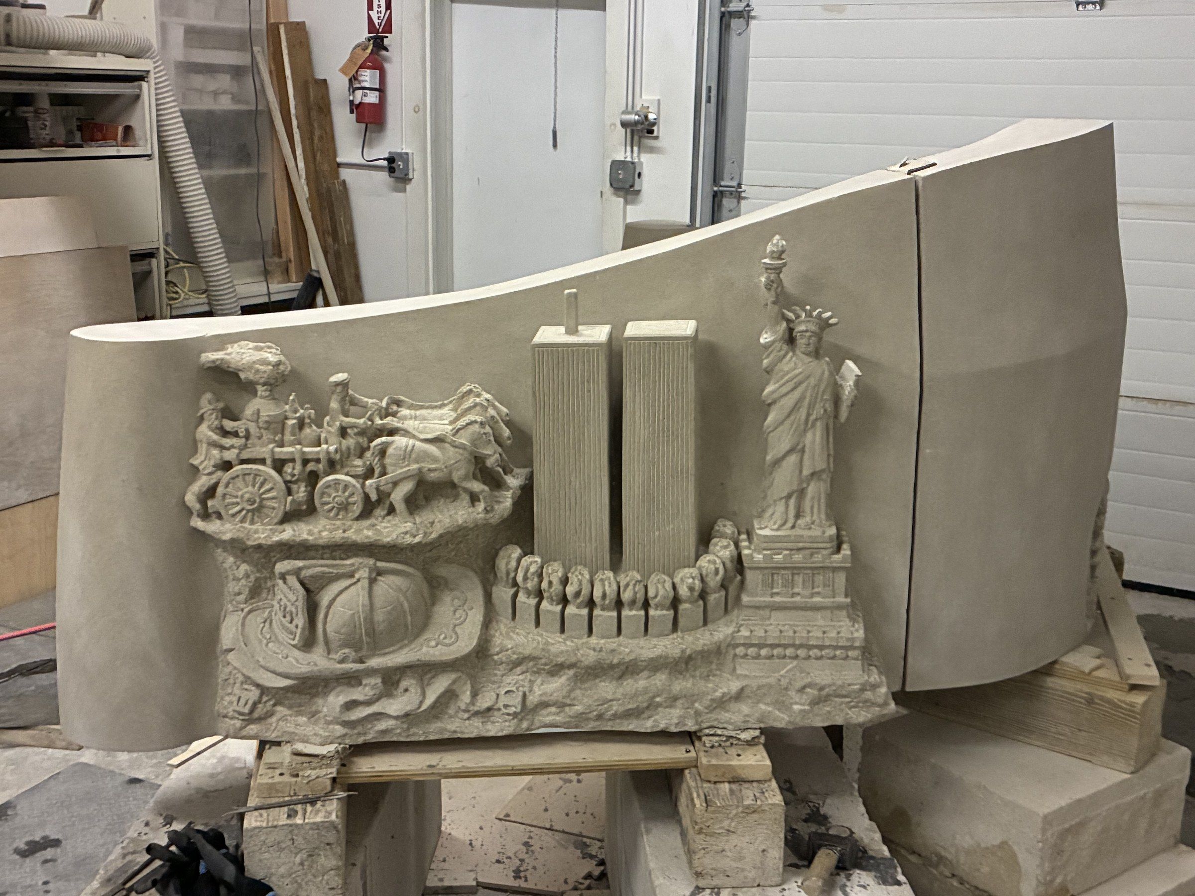 The Wyckoff Centennial Monument in Jean-Claude Marchionni's Midland Park studio, a curved three-block limestone relief raised on wooden trestles: at the centre, the Twin Towers rise straight above a row of eleven forms, half flame and half flower; at the left, a 19th-century horse-drawn fire engine with a fireman's helmet draped in the American flag at its base; at the right, the Statue of Liberty on her pedestal, torch raised. The whole piece unfolds in a single flowing wave.