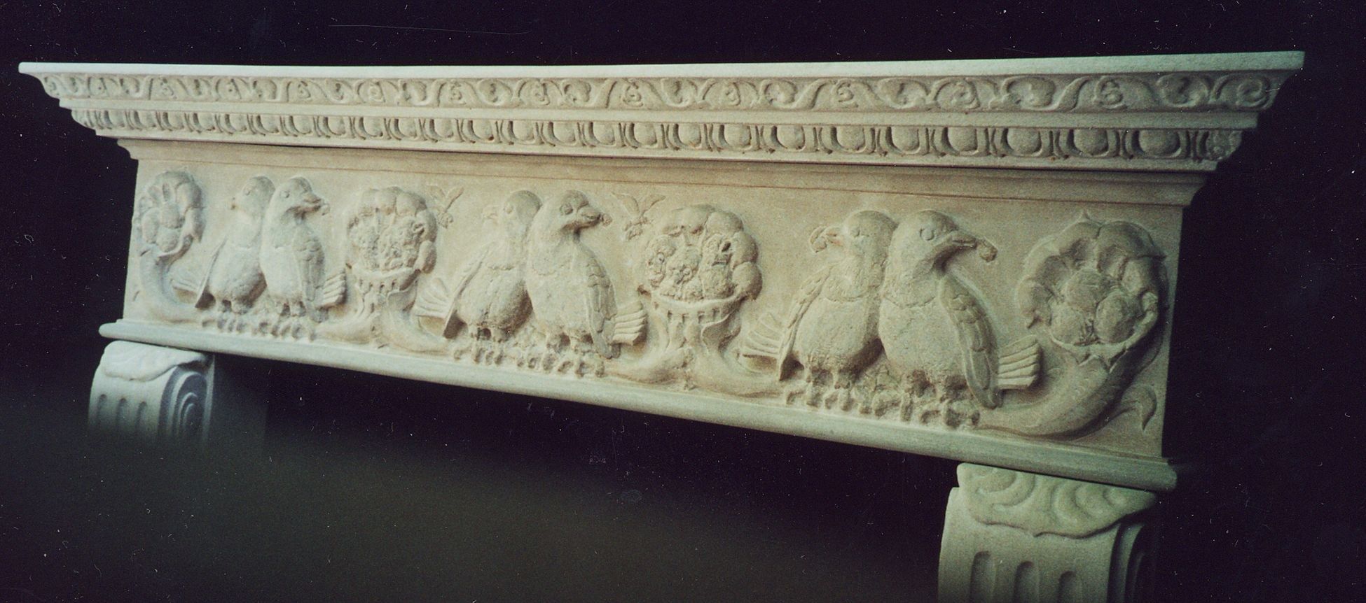 A carved limestone mantel photographed against a black backdrop: a bas-relief frieze of paired birds perched between baskets and cornucopias of fruit, beneath a cornice carved with a running scroll band over egg-and-dart, resting on fluted scroll consoles.