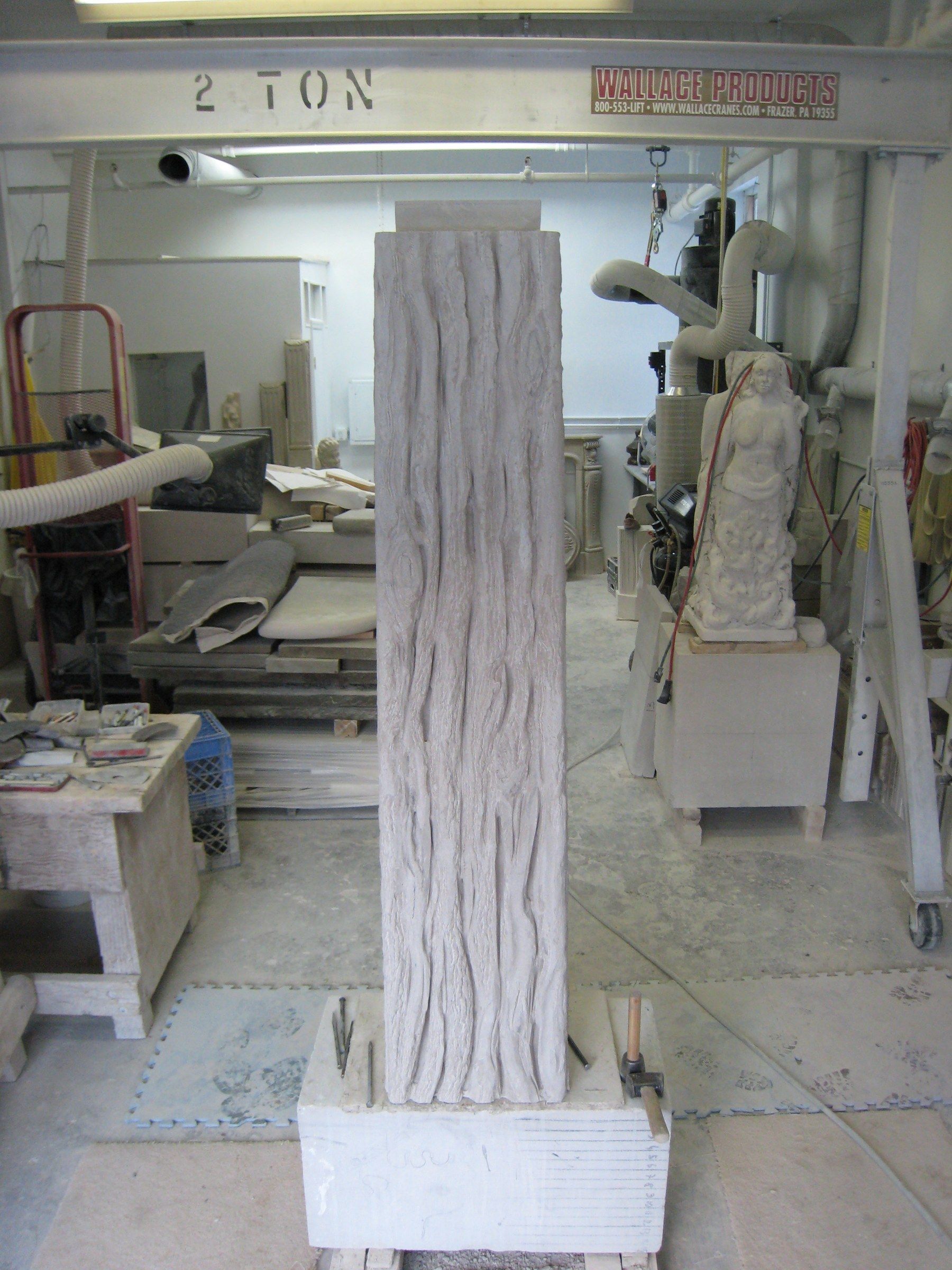The carved trunk column standing upright in the studio on a stone base, its surface cut with deep, wavering vertical grooves like bark.