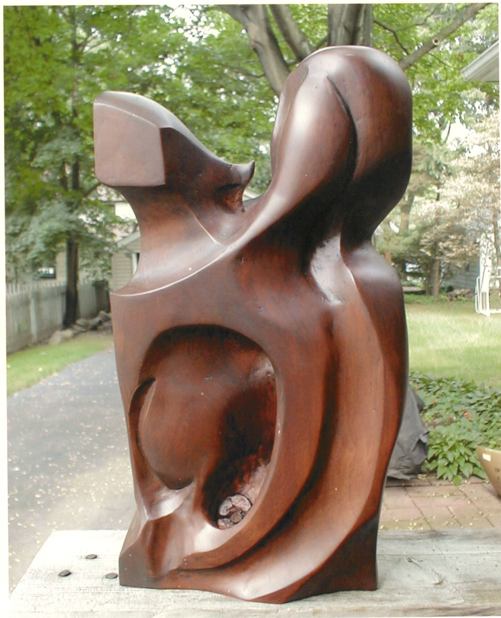 An abstract sculpture in dark, polished reddish-brown cherry wood, photographed outdoors on a wooden ledge: two upright forms rise from a single block, a deep oval hollow opens through the body below, and a small natural knot is left visible inside the hollow.