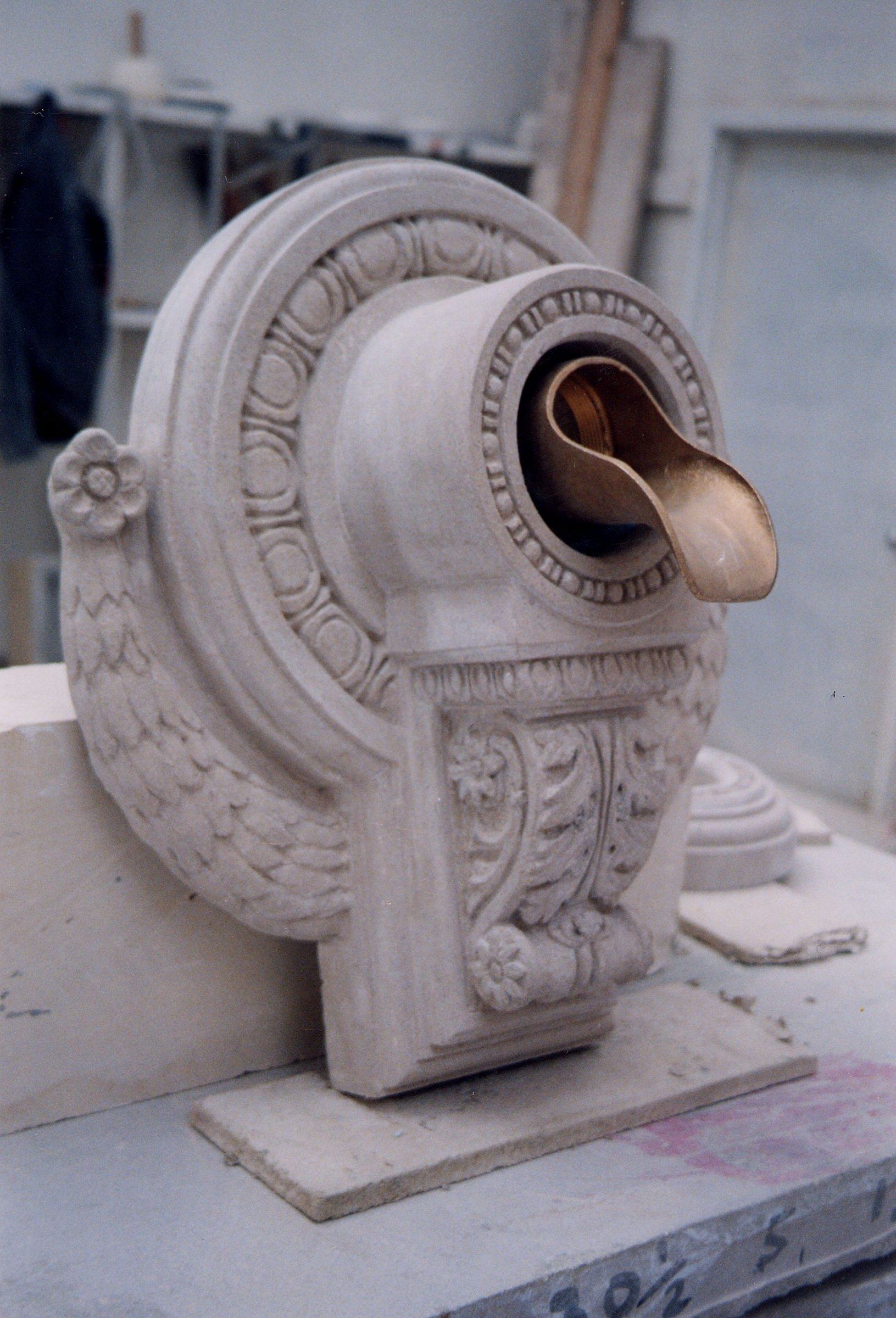 Three-quarter view of the limestone scupper on a workshop bench: a round back plate ringed with an egg-and-dart band, a projecting spout boss edged with bead-and-reel holding the curved bronze lip, an acanthus-scroll console with rosettes beneath, and a garland of overlapping leaves sweeping around one side to a carved rosette.