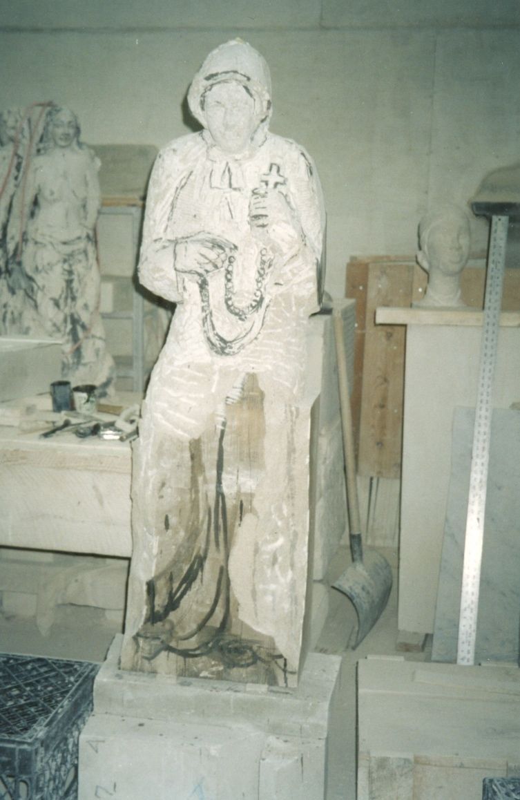 The statue roughed out in the workshop: the head, bonnet, hands, and rosary already defined, while the lower gown is still blocked in, with charcoal lines marking the folds still to be carved.